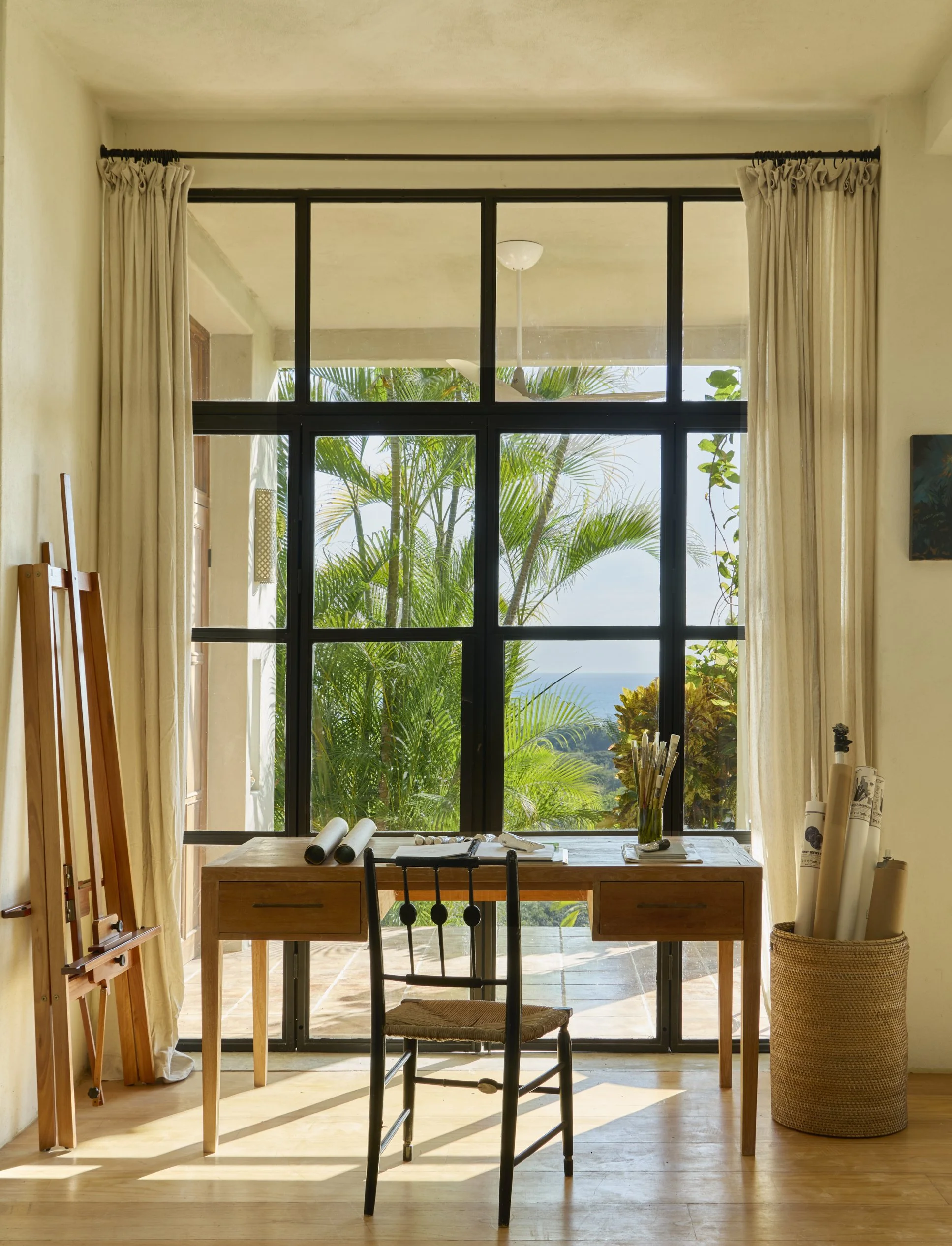 Interior room with a wooden desk and chair facing large window with curtains, overlooking lush green trees and a glimpse of the ocean in the distance.