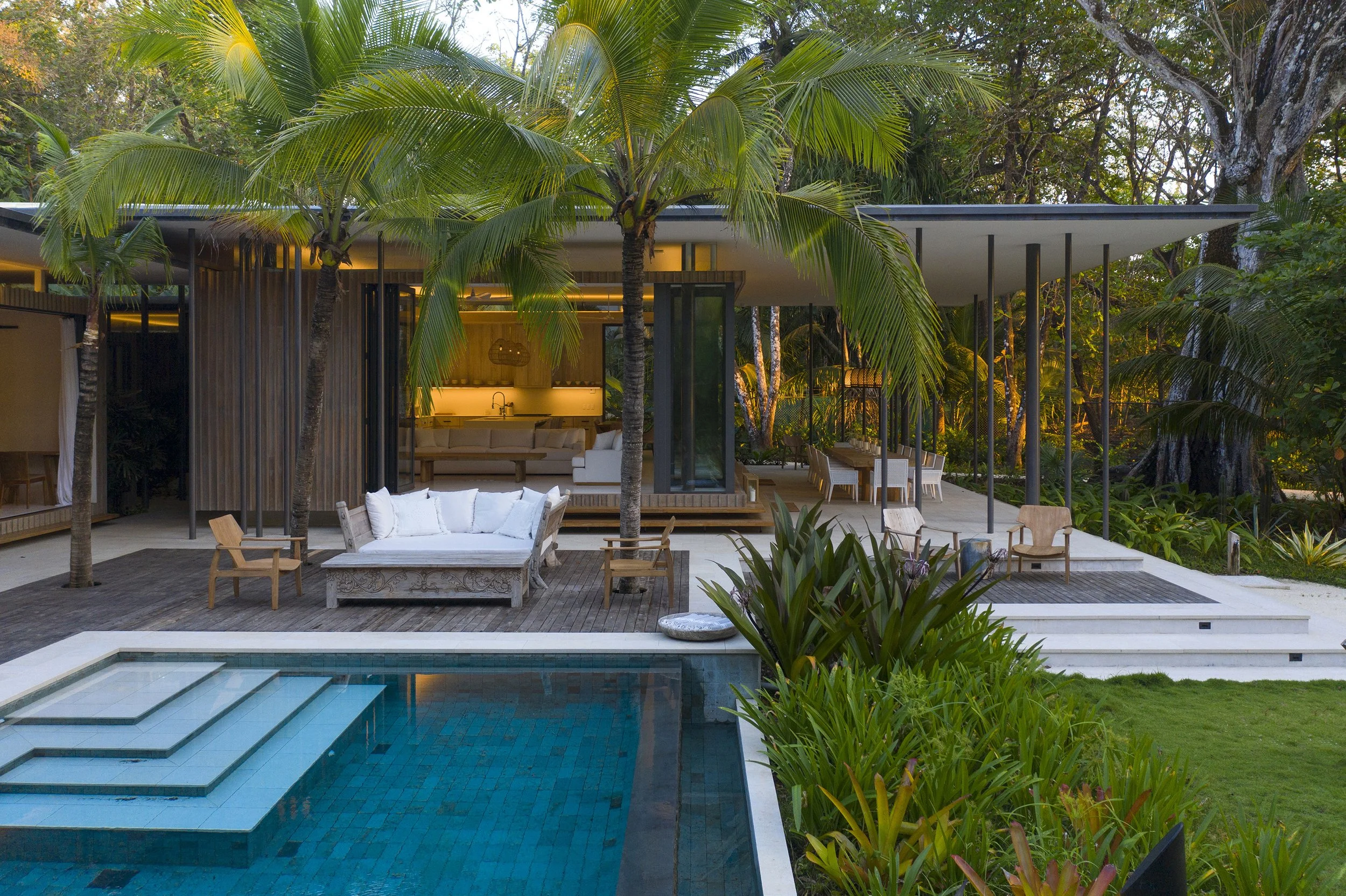 Modern house with outdoor furniture, palm trees, lush greenery, and a swimming pool in a tropical setting.