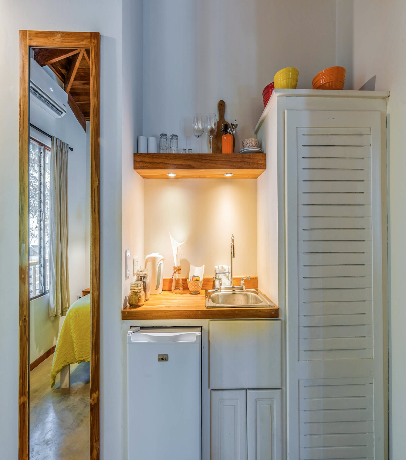 Small kitchen area with a wooden countertop, mini fridge, and open shelf with colorful bowls. Reflection in a large mirror shows a bedroom with a bed and window.