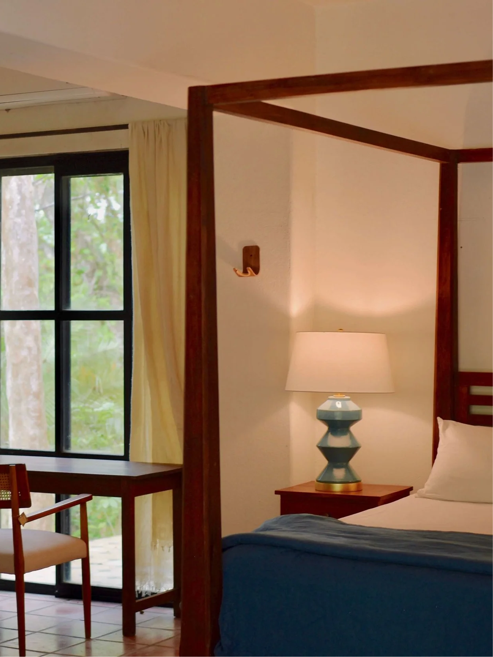 A bedroom with a bed, a bedside lamp, a wooden canopy bed frame, a window with white curtains, and outdoor greenery visible through the window.