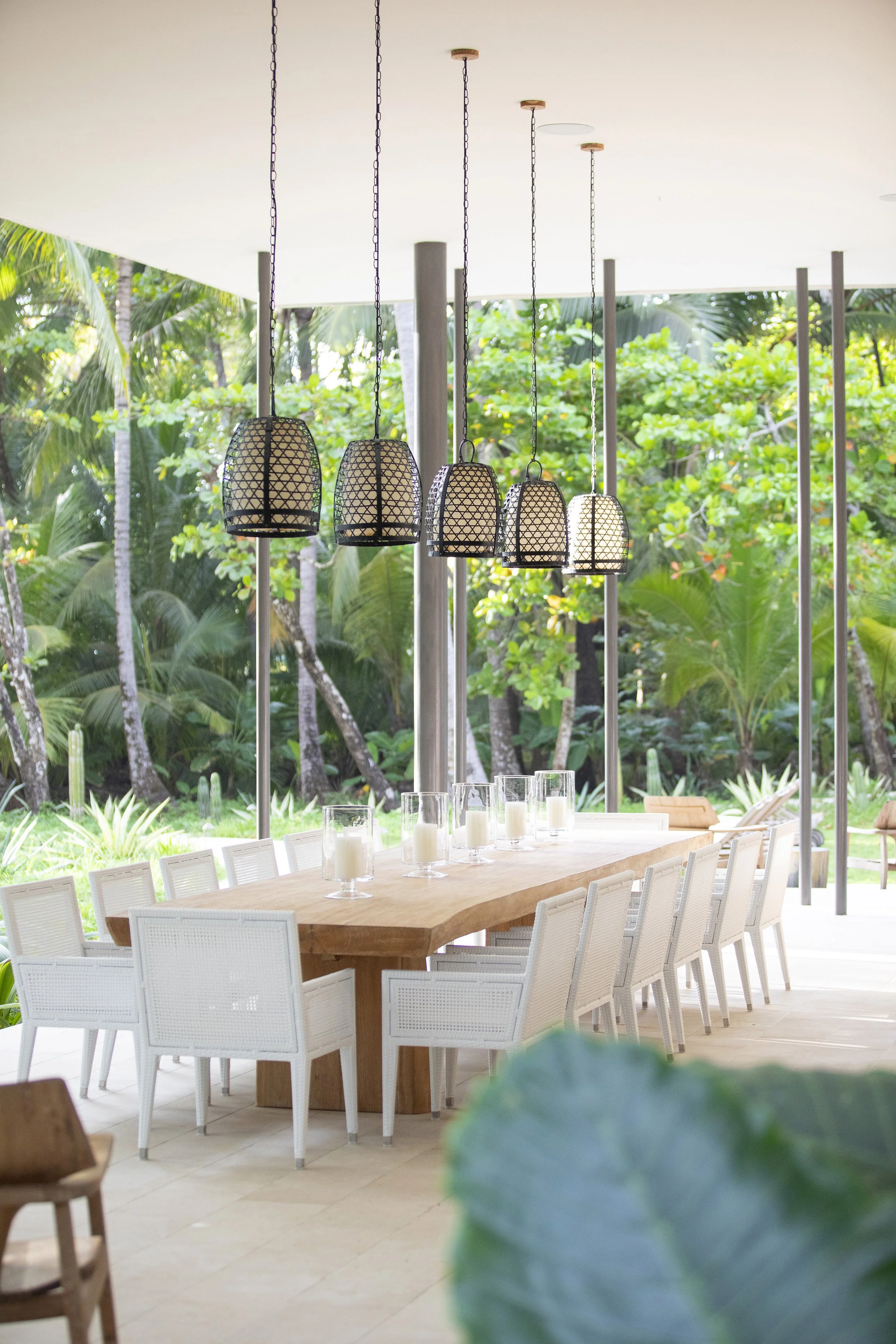 Outdoor dining area with a long wooden table, surrounded by white chairs, decorated with candles in glass holders. Overhead are five hanging lantern-style light fixtures, and in the background, lush green tropical plants and trees.