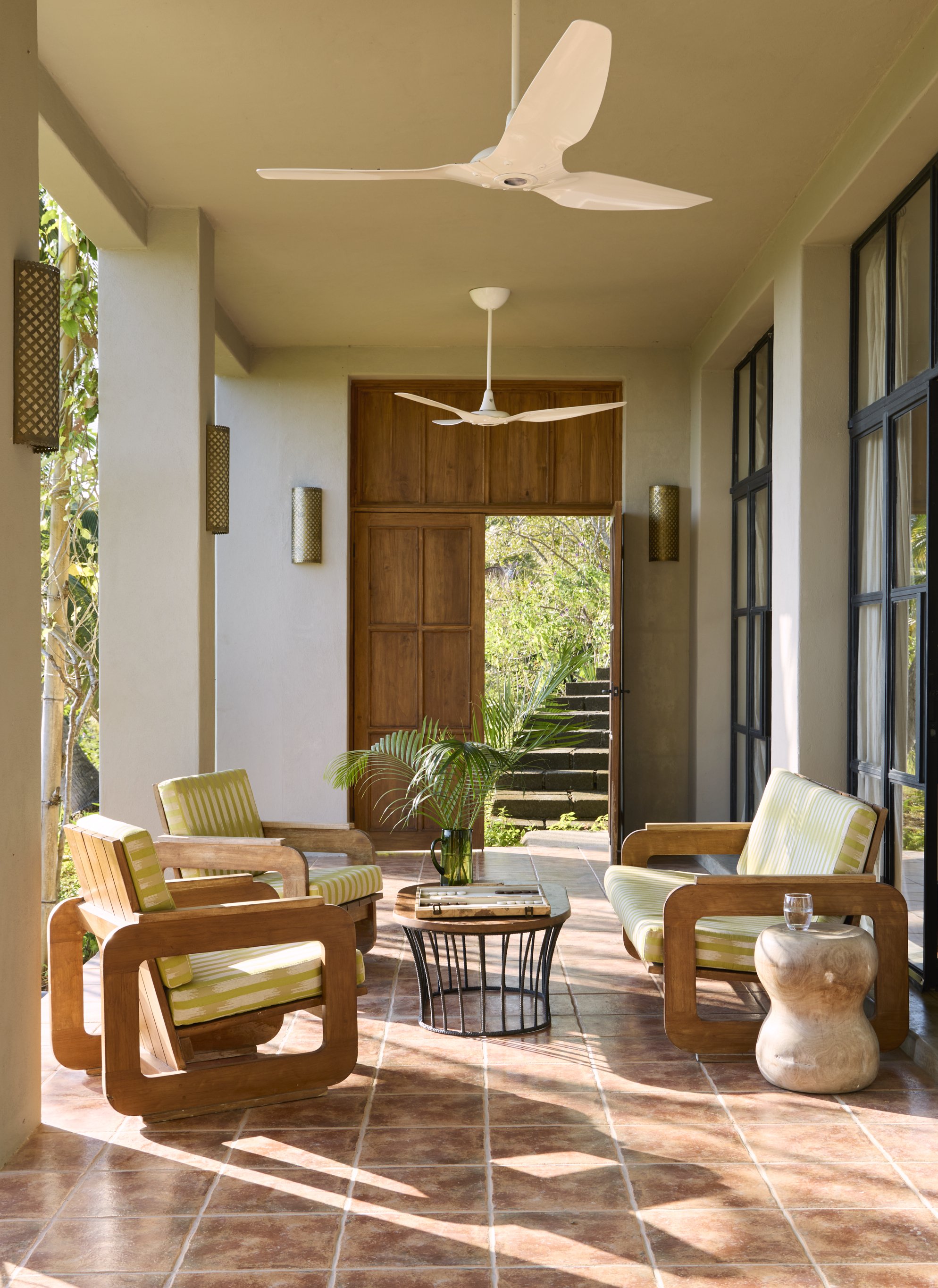 A porch with tiled floor, tropical plants, glass cup, and three wooden armchairs with yellow and white striped cushions, a small black side table, a green plant in a vase, an open door, and a ceiling fan
