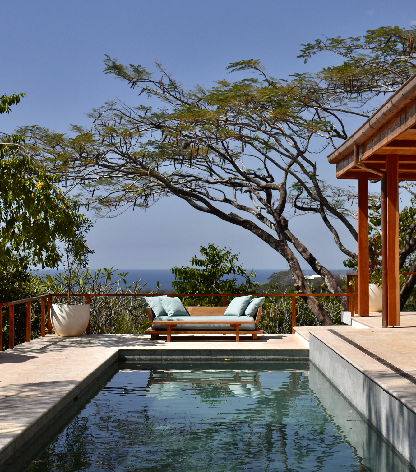 A luxurious outdoor patio with a rectangular swimming pool, a wooden lounge chair with cushions, and a large potted plant. There are trees and a clear blue sky in the background, with a view of the ocean.