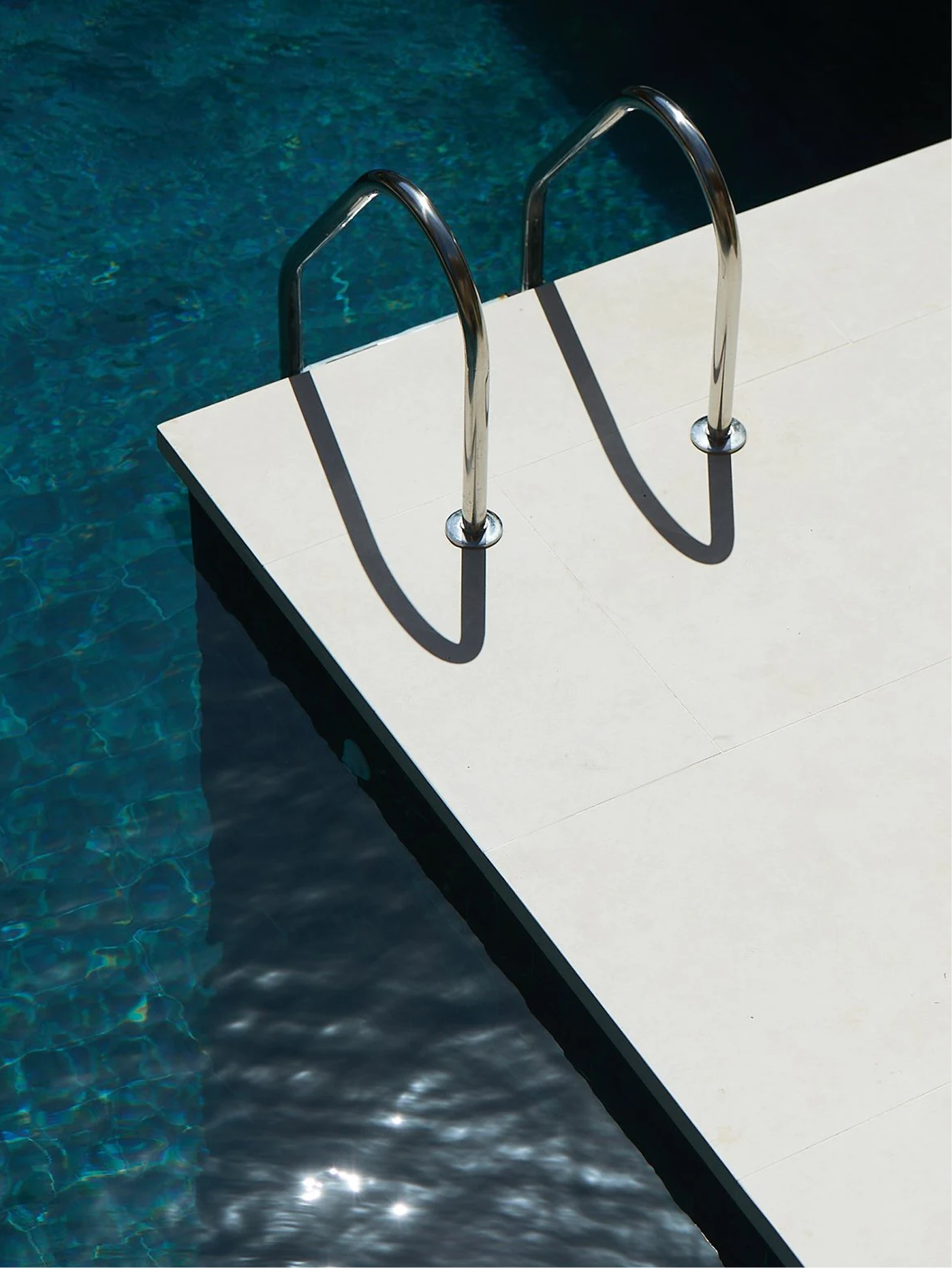 A metal pool ladder with two steps over a white tiled pool deck, leading into a dark blue swimming pool.