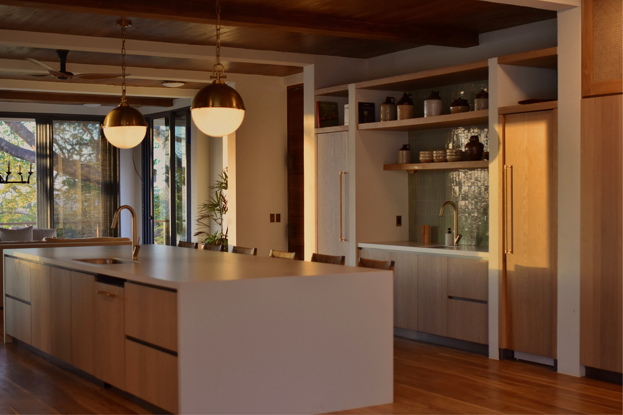 Modern kitchen with wooden cabinets, open shelves with jars and bowls, large island with a handle, pendant lights, and large windows showing a tree outside.