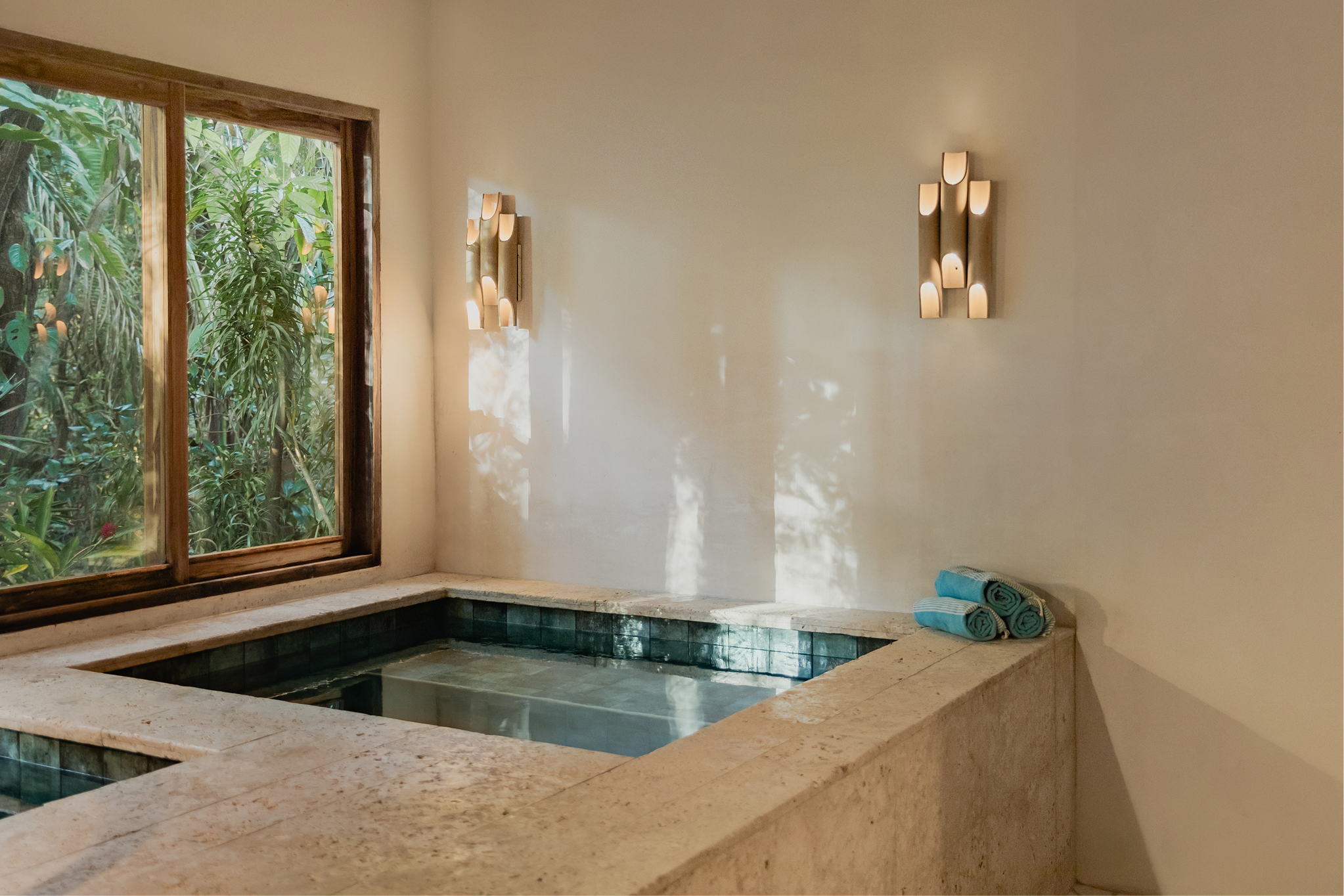 Indoor bathroom with a built-in bathtub next to a window with wooden frame, beige walls, candles on the wall, and folded towels on the corner of the bathtub.