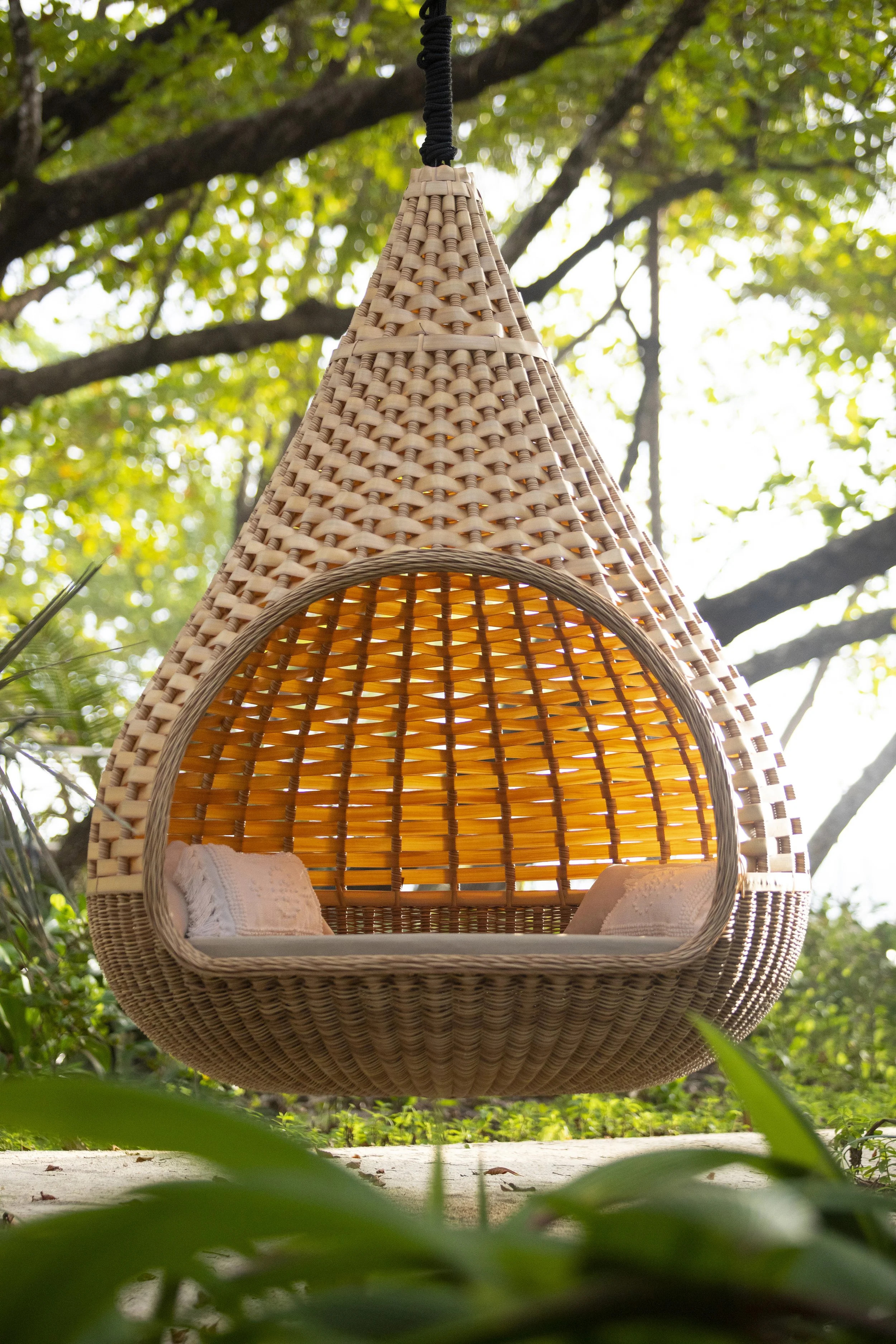 A hanging wicker chair with a rounded top and open front, containing cushions inside, suspended from a tree branch amidst green foliage and sunlight filtering through the leaves.