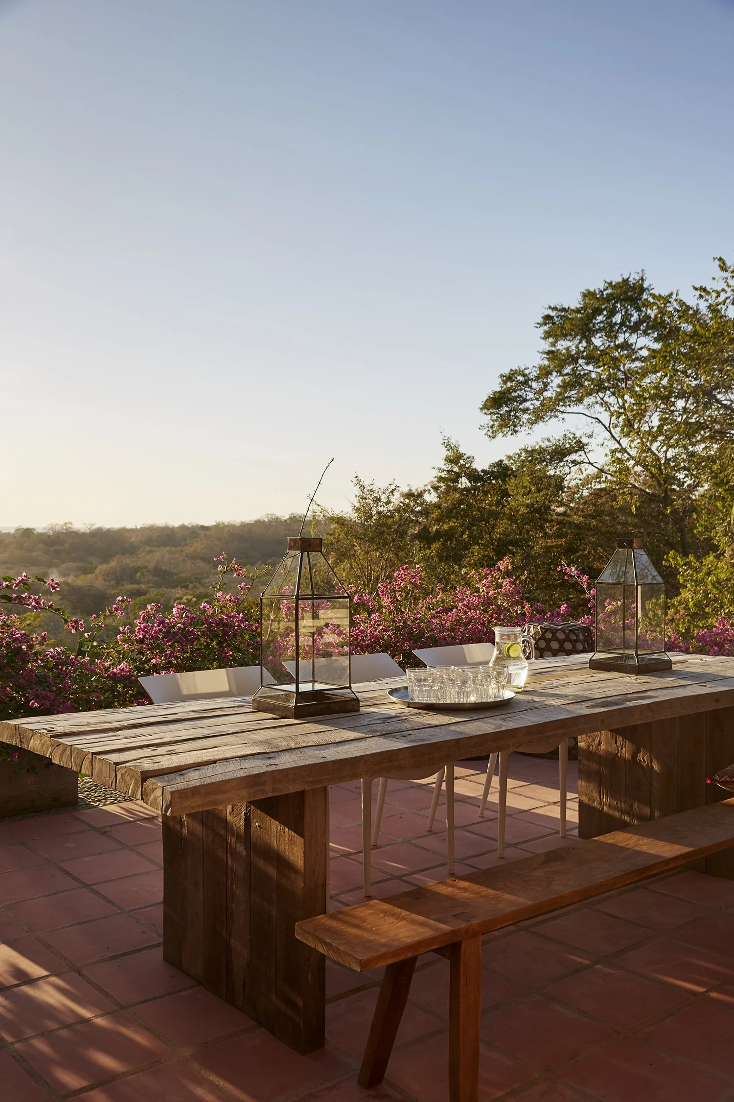 Outdoor wooden dining table with lanterns, glasses, and a tray with lemon slices, surrounded by pink flowering plants and trees in the distance during sunset.