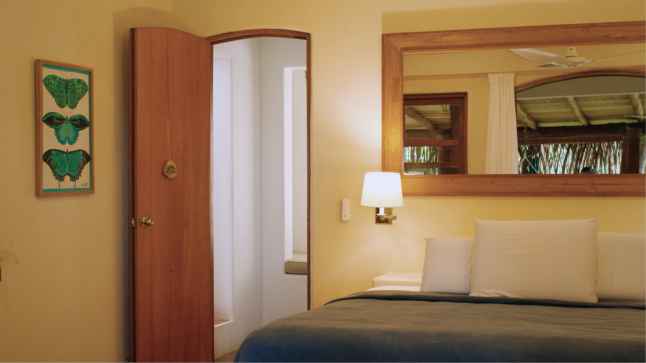 A bedroom featuring a bed with white pillows and a dark blanket, a wooden framed mirror on the wall, a wall-mounted lamp, a partially open wooden door, and a framed butterfly artwork on the yellow wall.