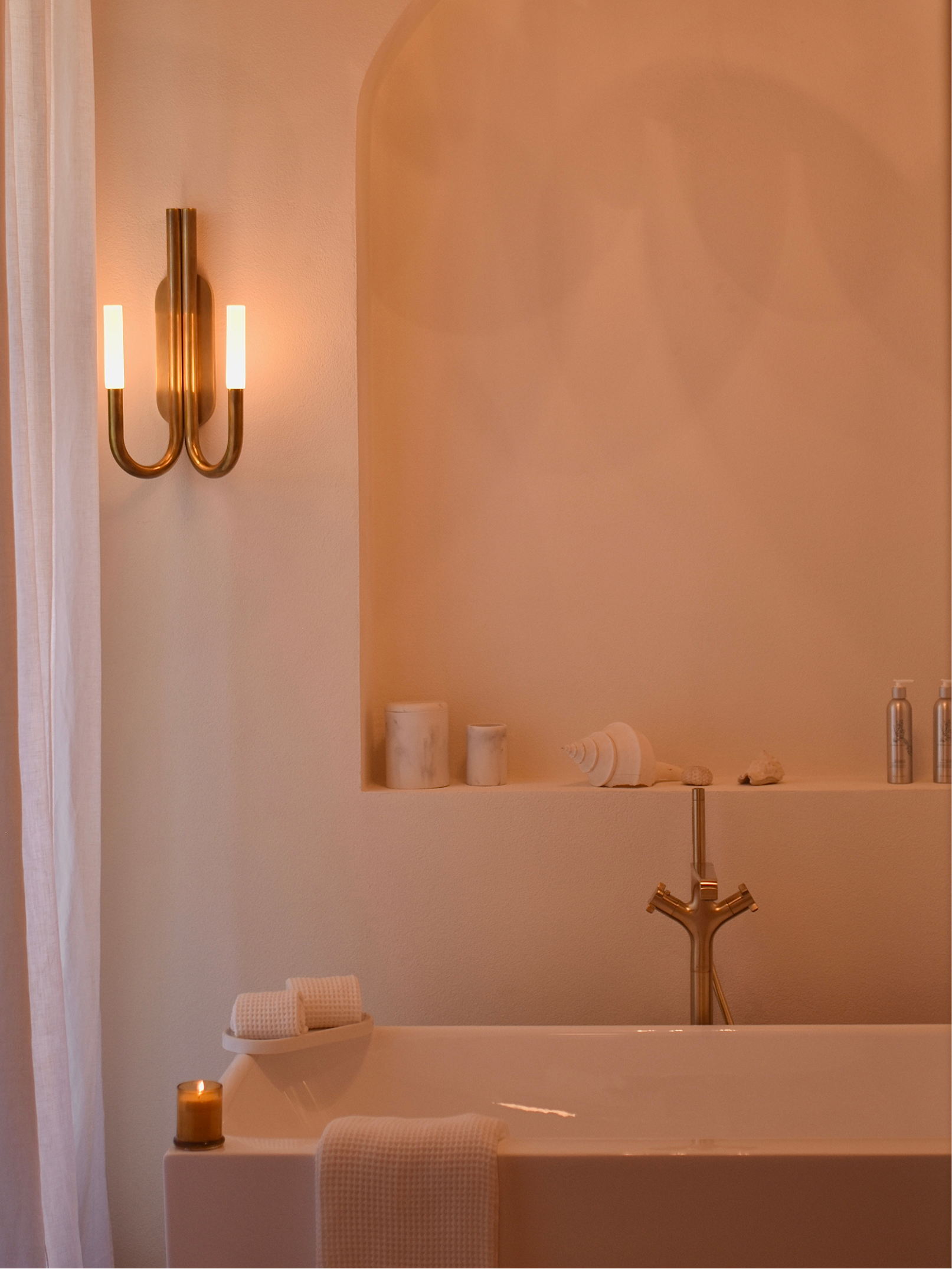 A bathroom with a bathtub, gold-colored faucet, and a towel. The wall has a ledge with decorative items including candles, shells, and stones. There is a gold-colored wall sconce with two lights, and a white shower curtain partially visible on the left.