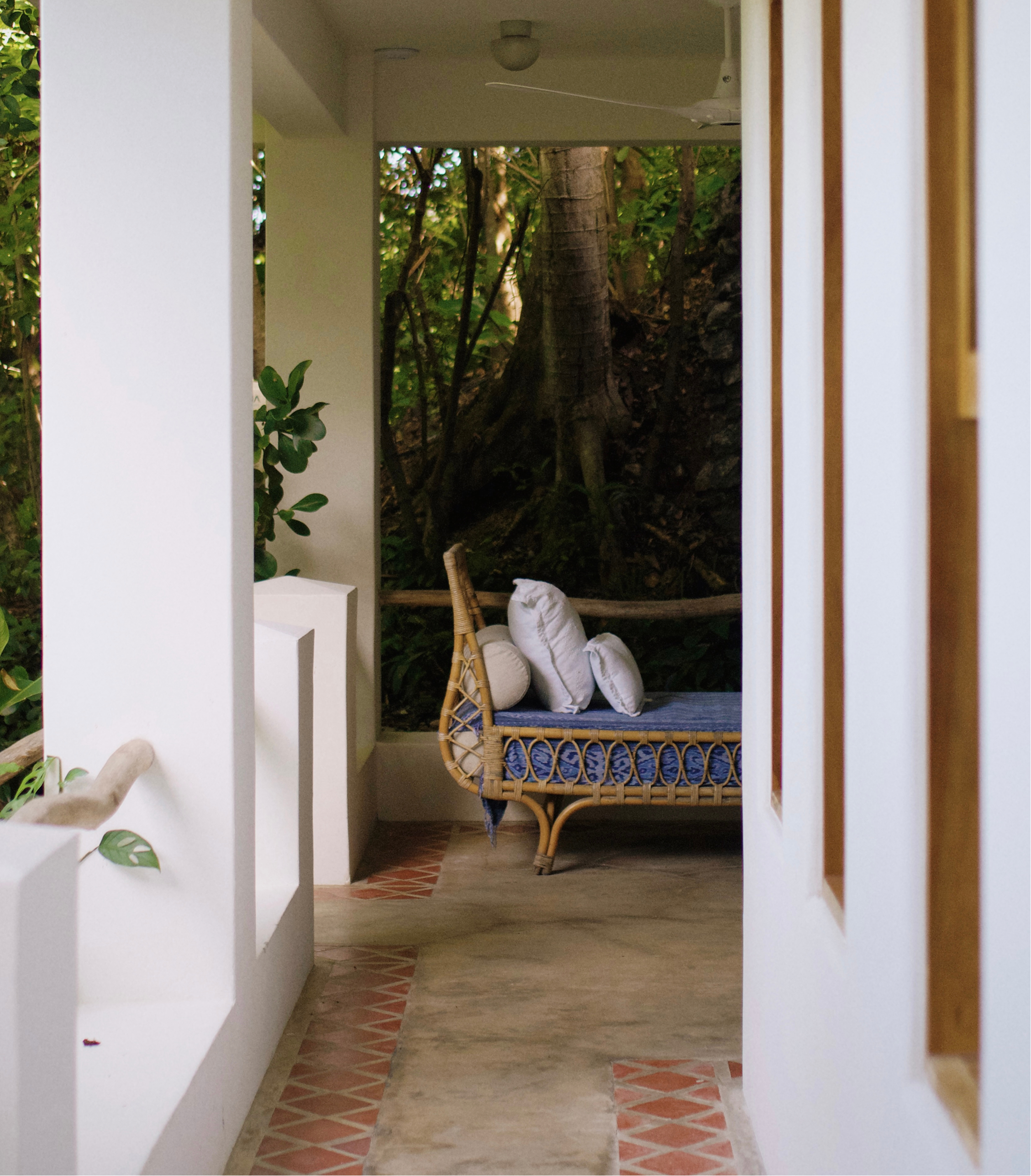 View of an outdoor patio area through a narrow walkway, featuring a wicker bench with white cushions and surrounded by greenery, with a large tree in the background.