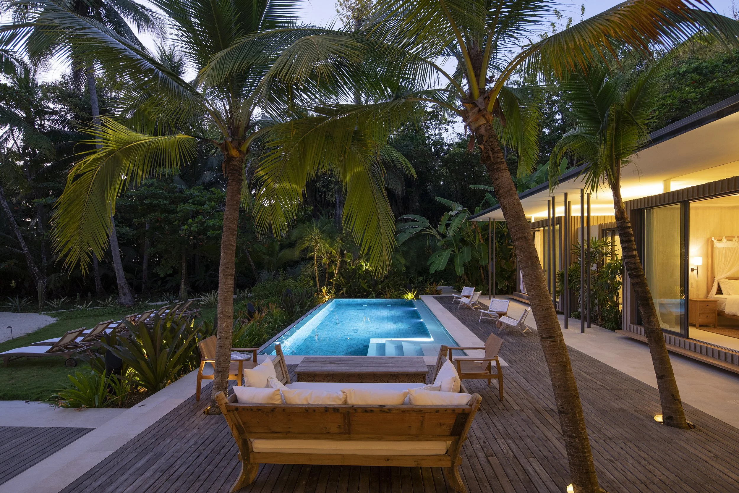 A modern backyard with a swimming pool, surrounded by palm trees and lounge chairs, with a contemporary house illuminated at dusk.