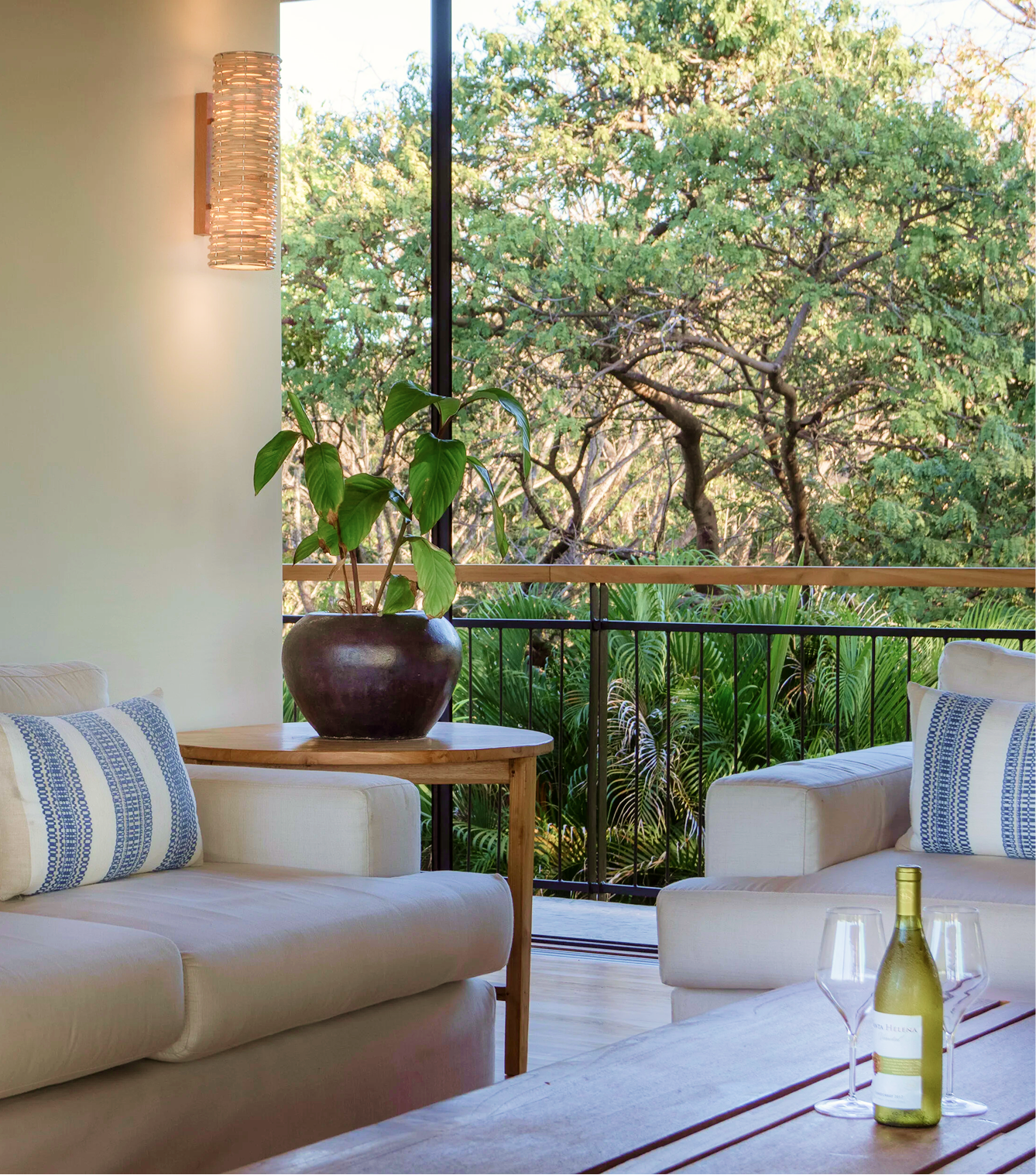 Outdoor patio with white cushioned seating, a large potted plant, a wooden table, and a view of green trees through a large window or open balcony.
