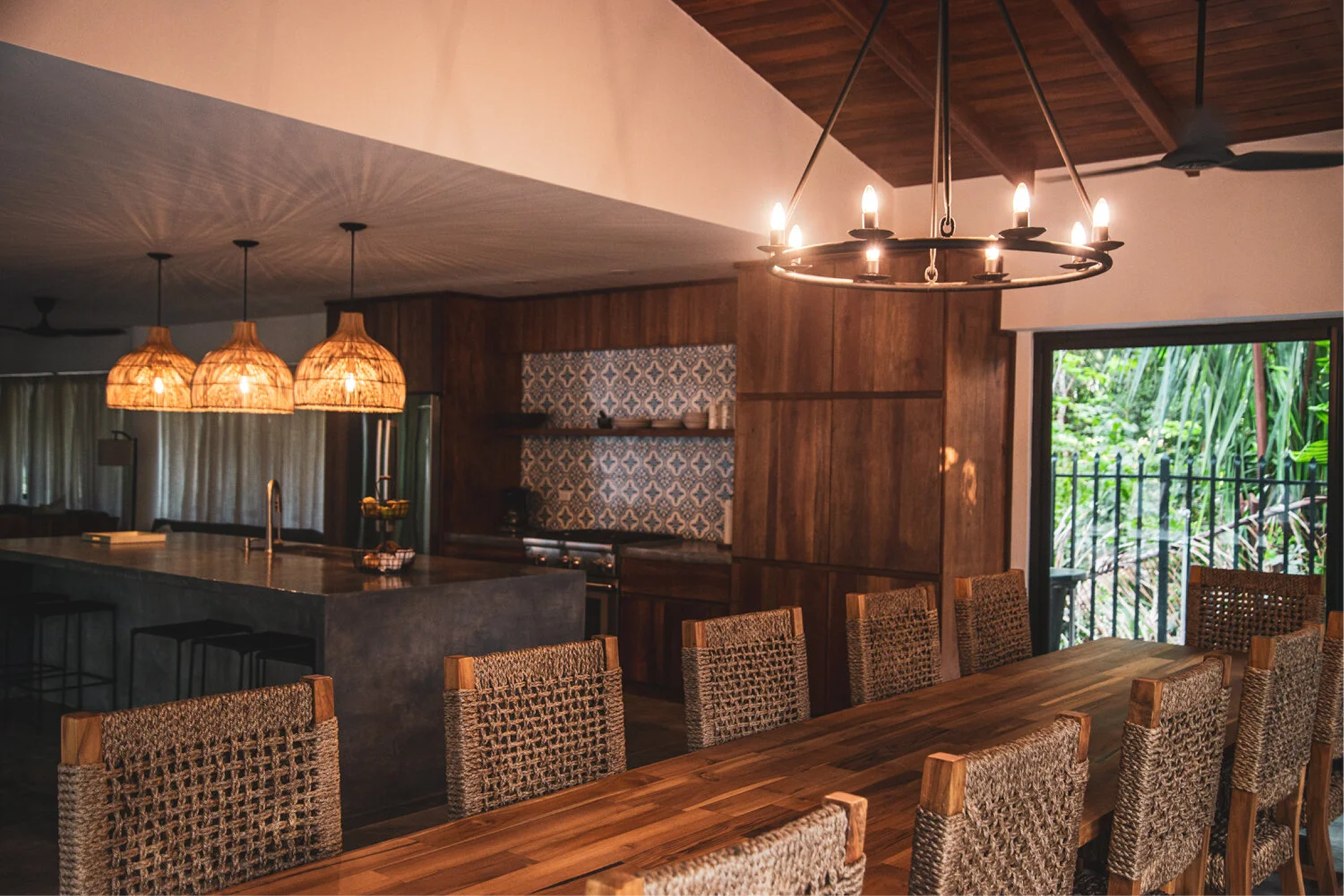 Interior of a modern kitchen and dining area with wooden walls and ceiling, wicker chairs around a wooden dining table, hanging pendant lights above the kitchen island, a chandelier with lit candles, and an open sliding door showing green outdoor scenery.