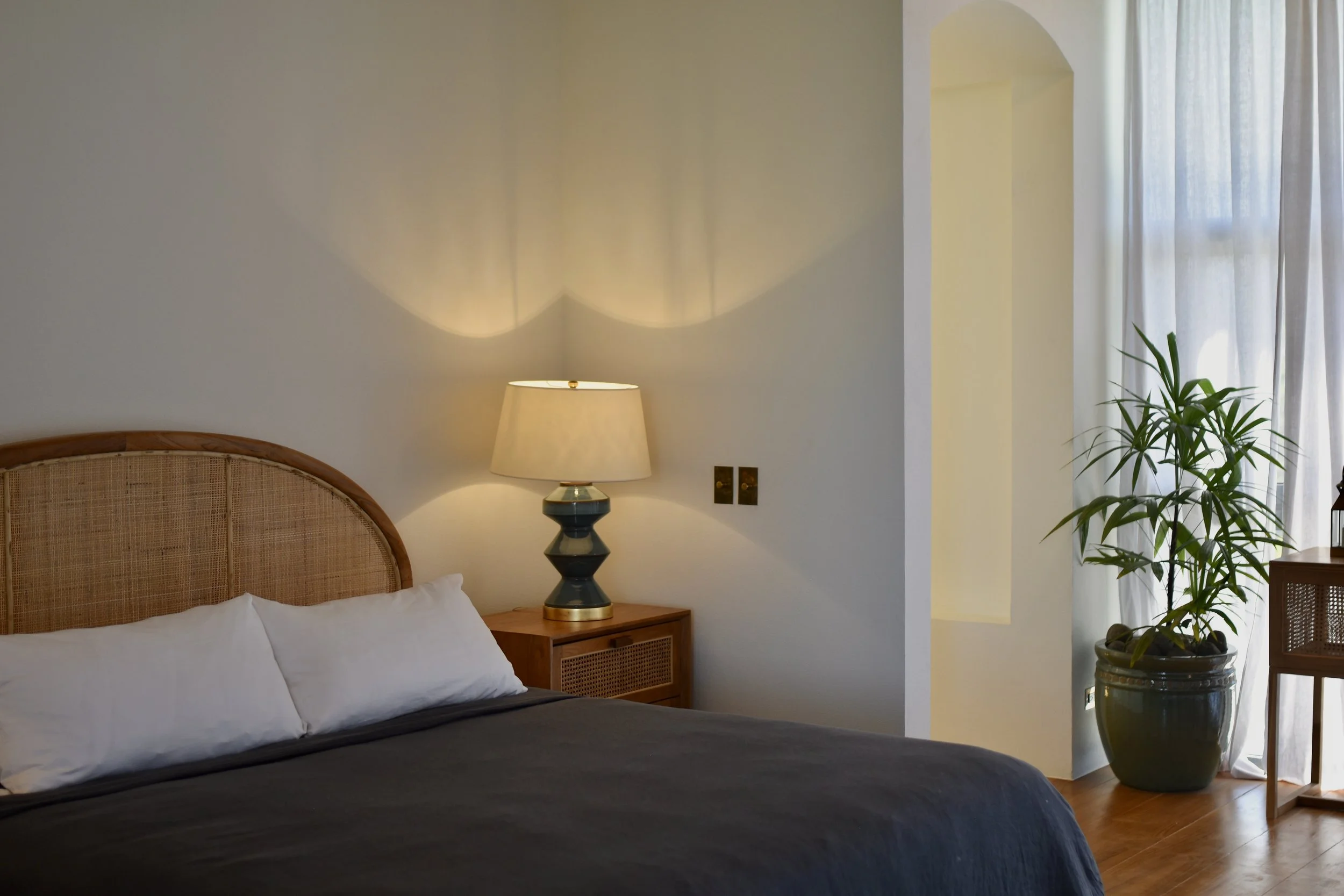 A bedroom with a wooden headboard, white bedding, a black bedspread, a nightstand with a modern lamp, a potted plant, and sheer curtains in the background.