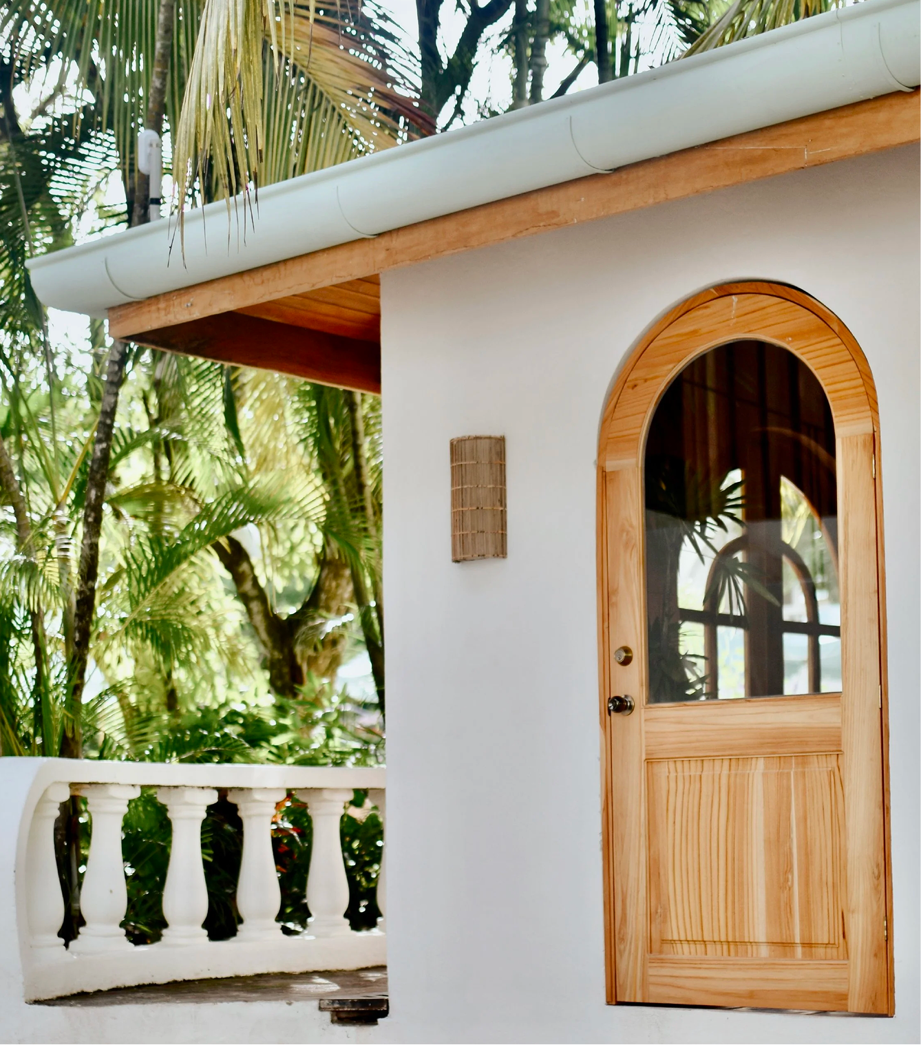A wooden door with an arched window in a white stucco wall, surrounded by lush green palm trees on a sunny day.