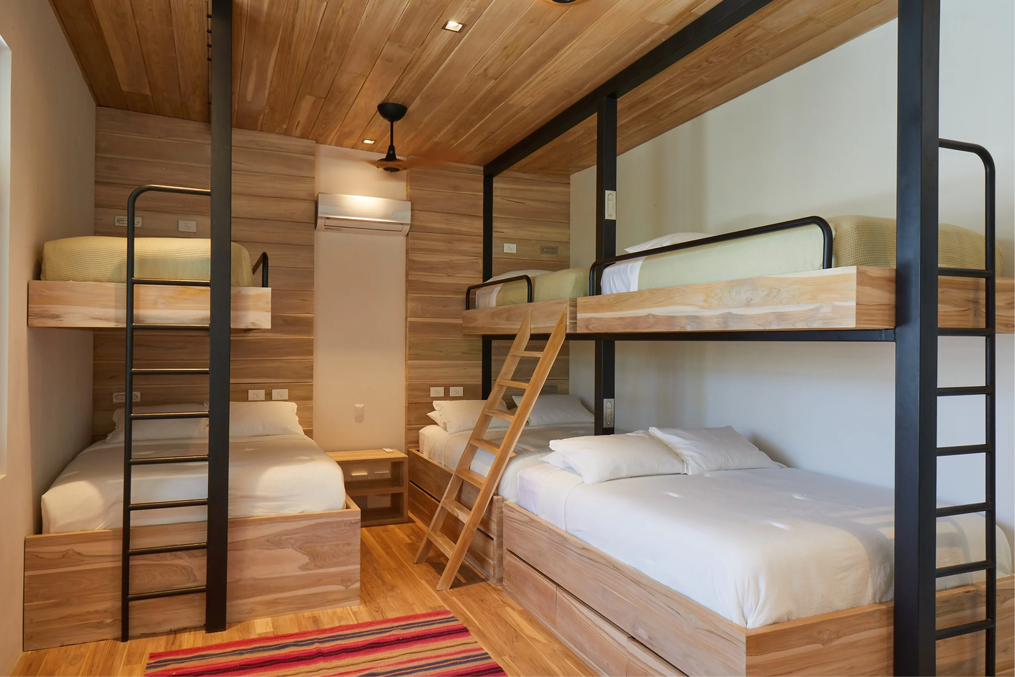 A room with two sets of bunk beds made of wood and metal, with mattresses and pillows. The room has a wooden ceiling and wall paneling, with a ceiling fan, air conditioning unit, and colorful rug on the floor.