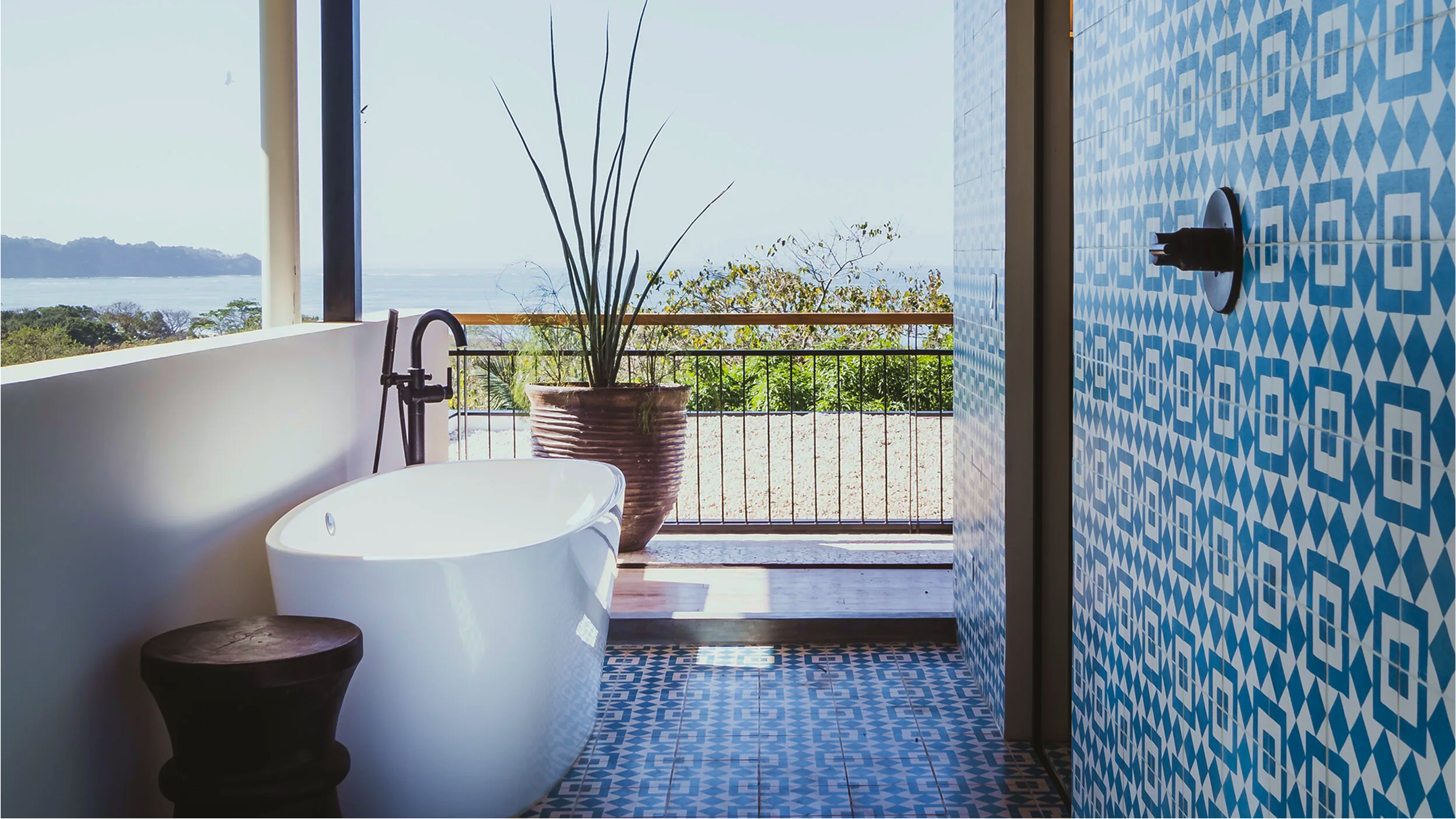 Bathtub with a view of the ocean and a balcony, decorated with a large potted plant and blue geometric patterned tiles.