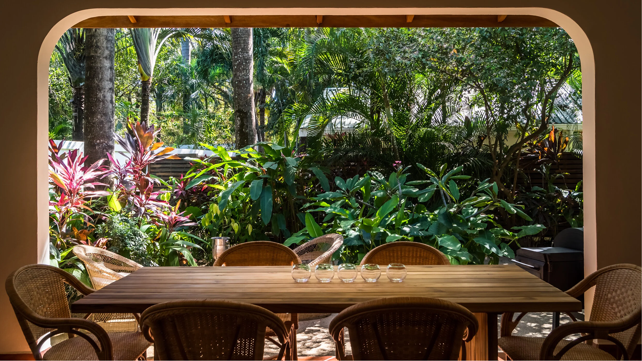 A view from inside an open-air dining area, looking out onto a lush tropical garden with various trees and plants.