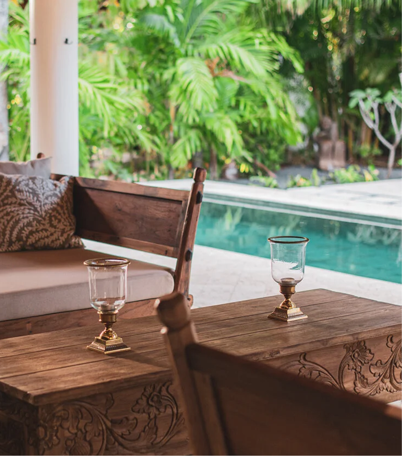 A cozy outdoor seating area with a wooden table, two glass candle holders, and a wooden chair with decorative carvings. In the background, there's a swimming pool and lush green tropical plants.