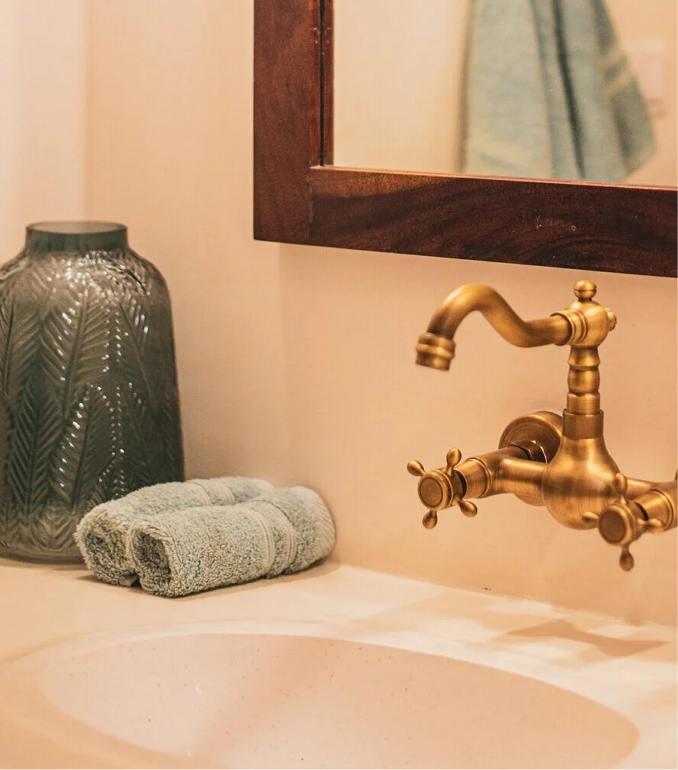 Decorative bathroom sink area with a rolled towel, a dark glass vase with patterns, a brass faucet, and a mirror with a wooden frame.