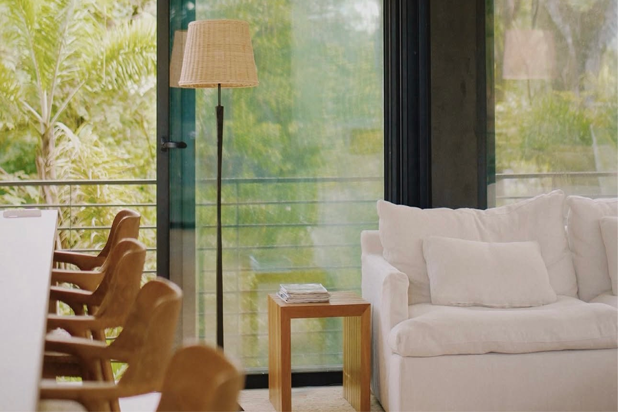 Interior view of a modern living room with a white couch, wooden side table with magazines, a window with black frame, glass door, and a stand lamp with a woven lampshade, overlooking green outdoor scenery.