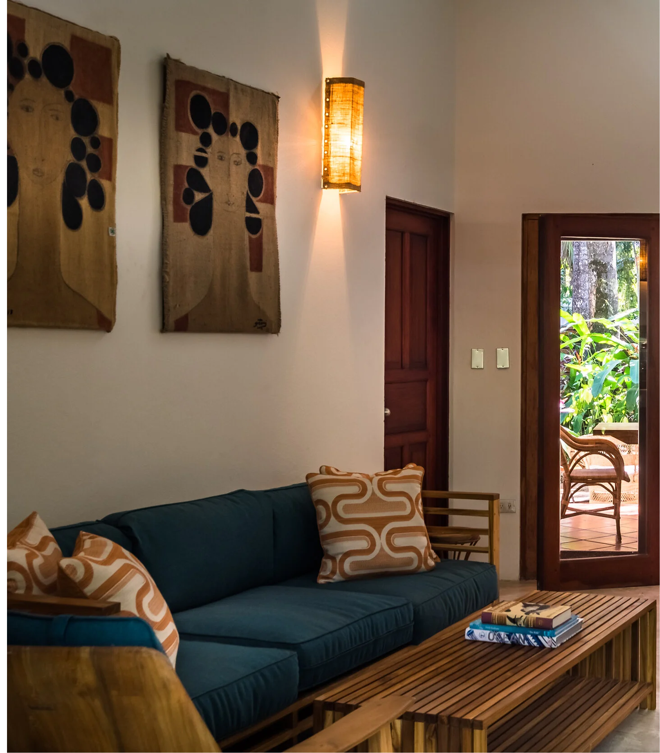 Living room with a blue couch, patterned throw pillows, wooden coffee table with books, wall art, wall-mounted lamp, open door leading to a patio with outdoor furniture, and greenery outside.