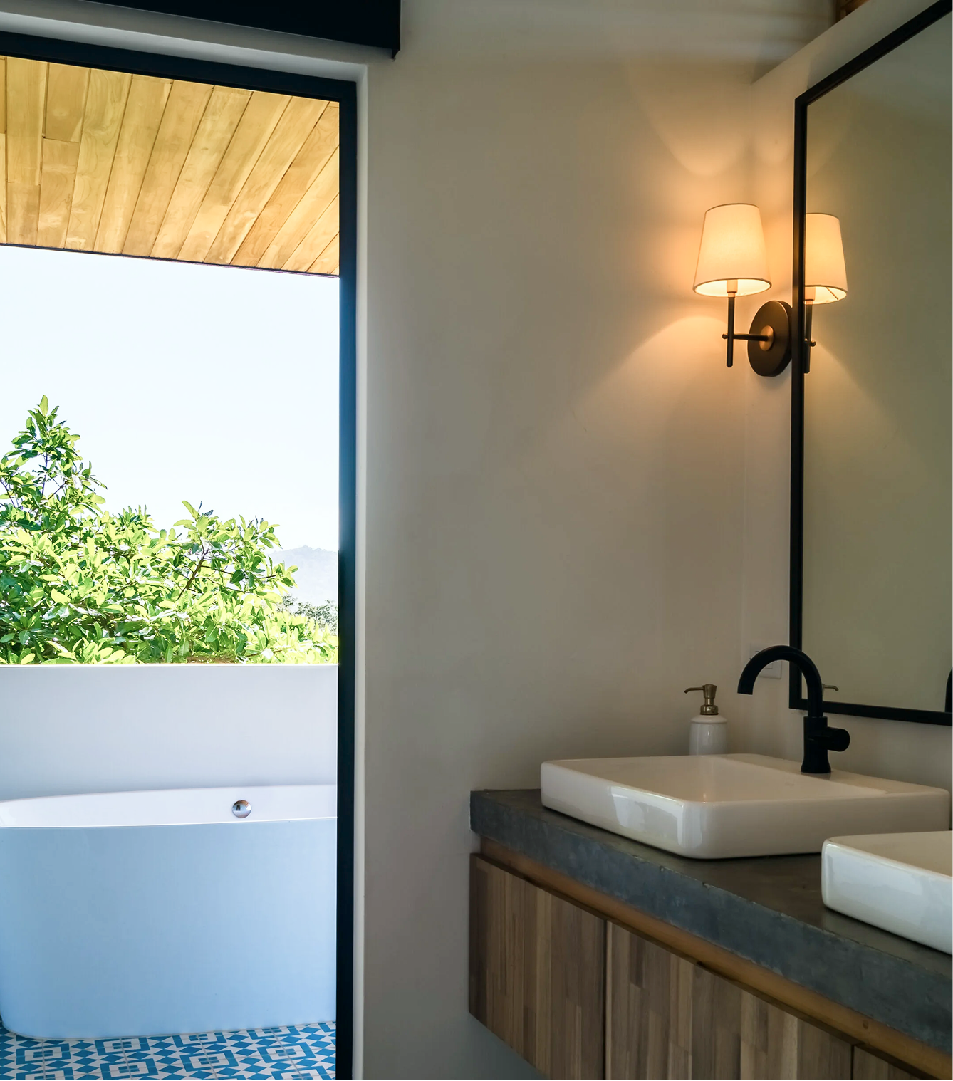 A bathroom with a white square sink, black faucet, wooden cabinet, wall-mounted lamp with shades, and a large mirror. Outside view through the open door shows greenery and a wooden ceiling.
