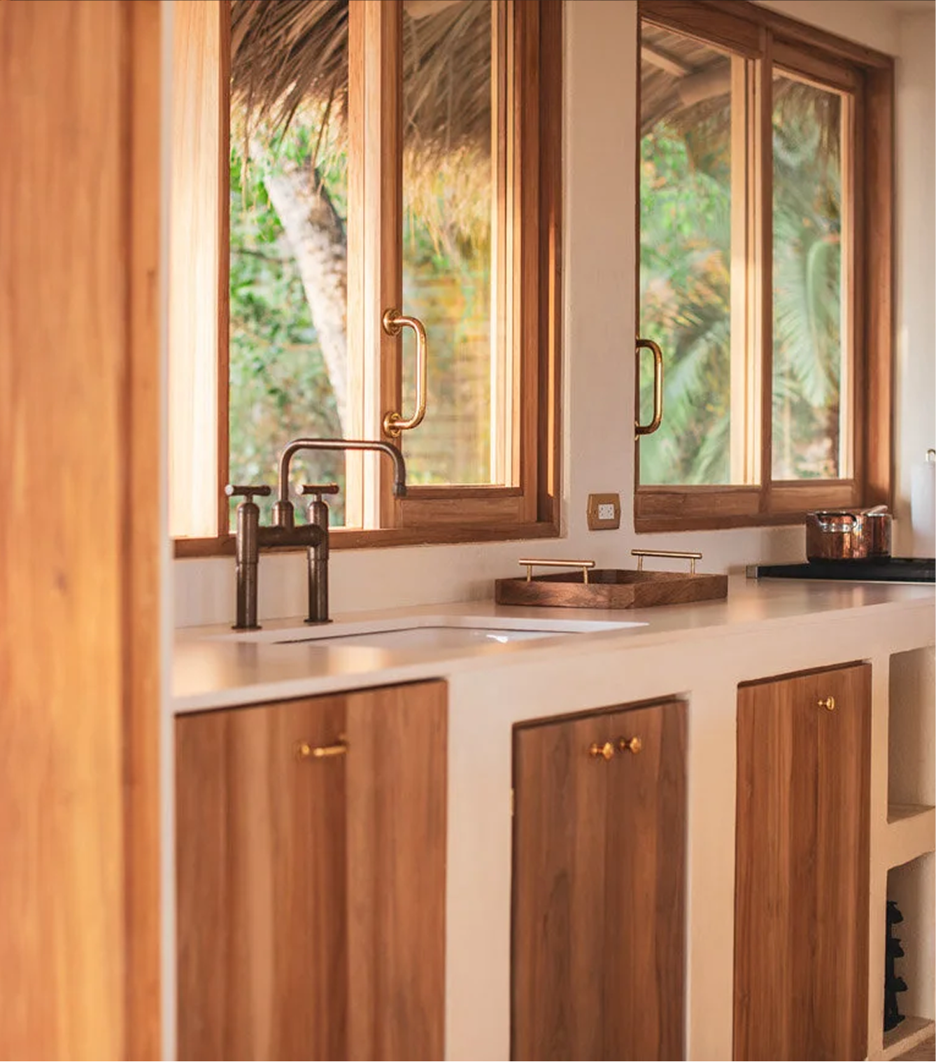 A kitchen with wooden cabinets and a cream-colored countertop. There are two windows with wooden frames showing green foliage outside. A black faucet is above a sink, and a copper pot is on the stove.