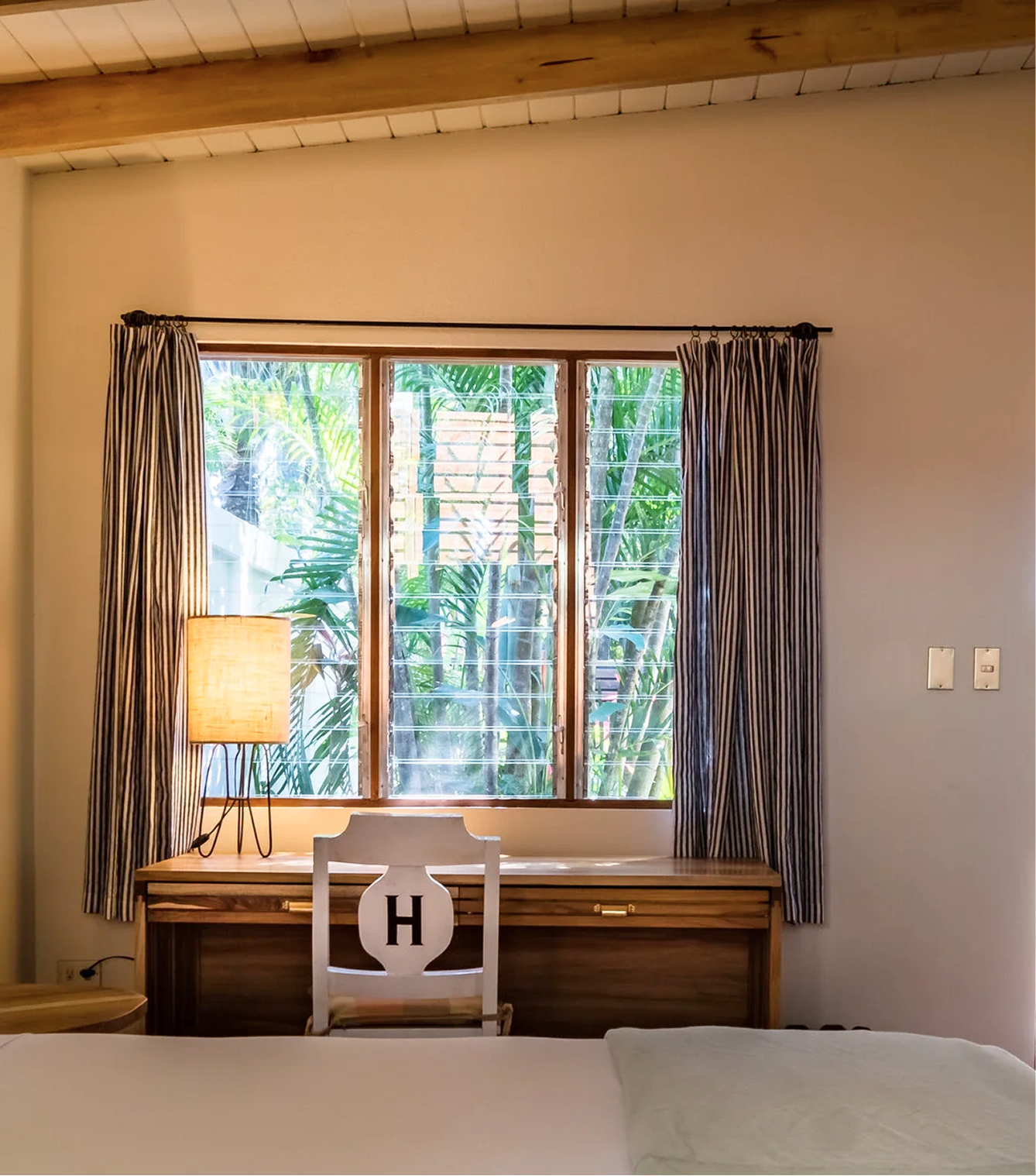 Bedroom with a wooden desk, a white chair with an 'H' on the back, a lamp, and a window with striped curtains showing green foliage outside.