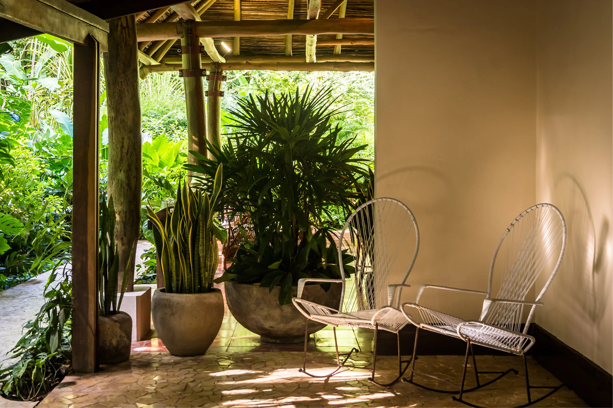 A shaded outdoor patio or porch area with potted plants, white metal chairs, and a lush green garden visible in the background.