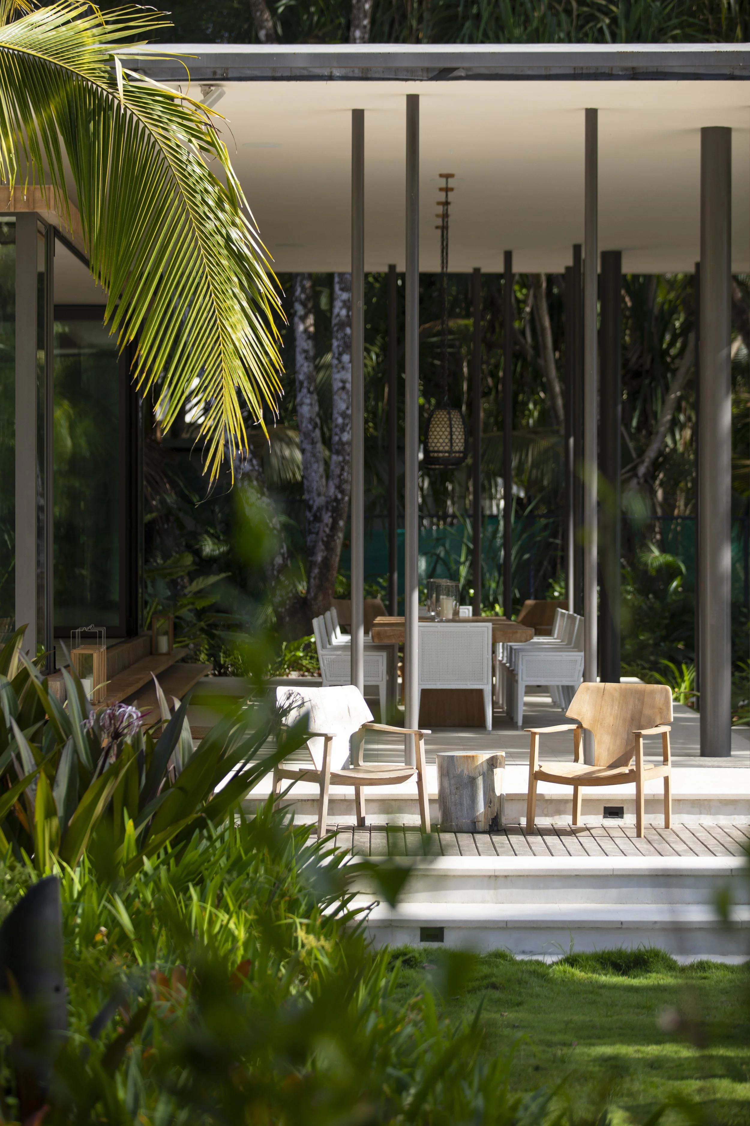 Outdoor seating area with wooden chairs and tables, surrounded by lush green plants, including palm leaves, with a modern house in the background featuring large glass windows and a white ceiling.
