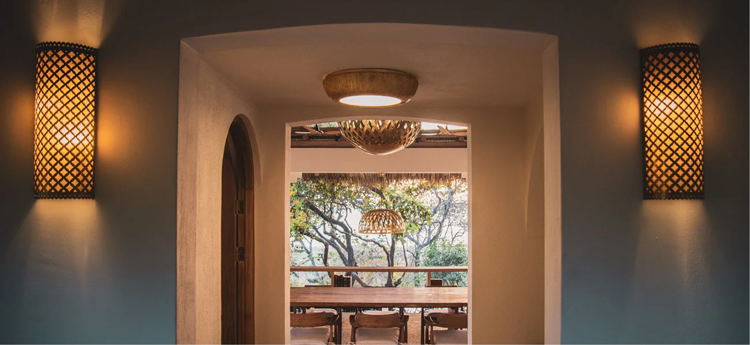 Interior corridor leading to an outdoor dining area with large wooden table, surrounded by chairs, overlooking trees and a balcony.