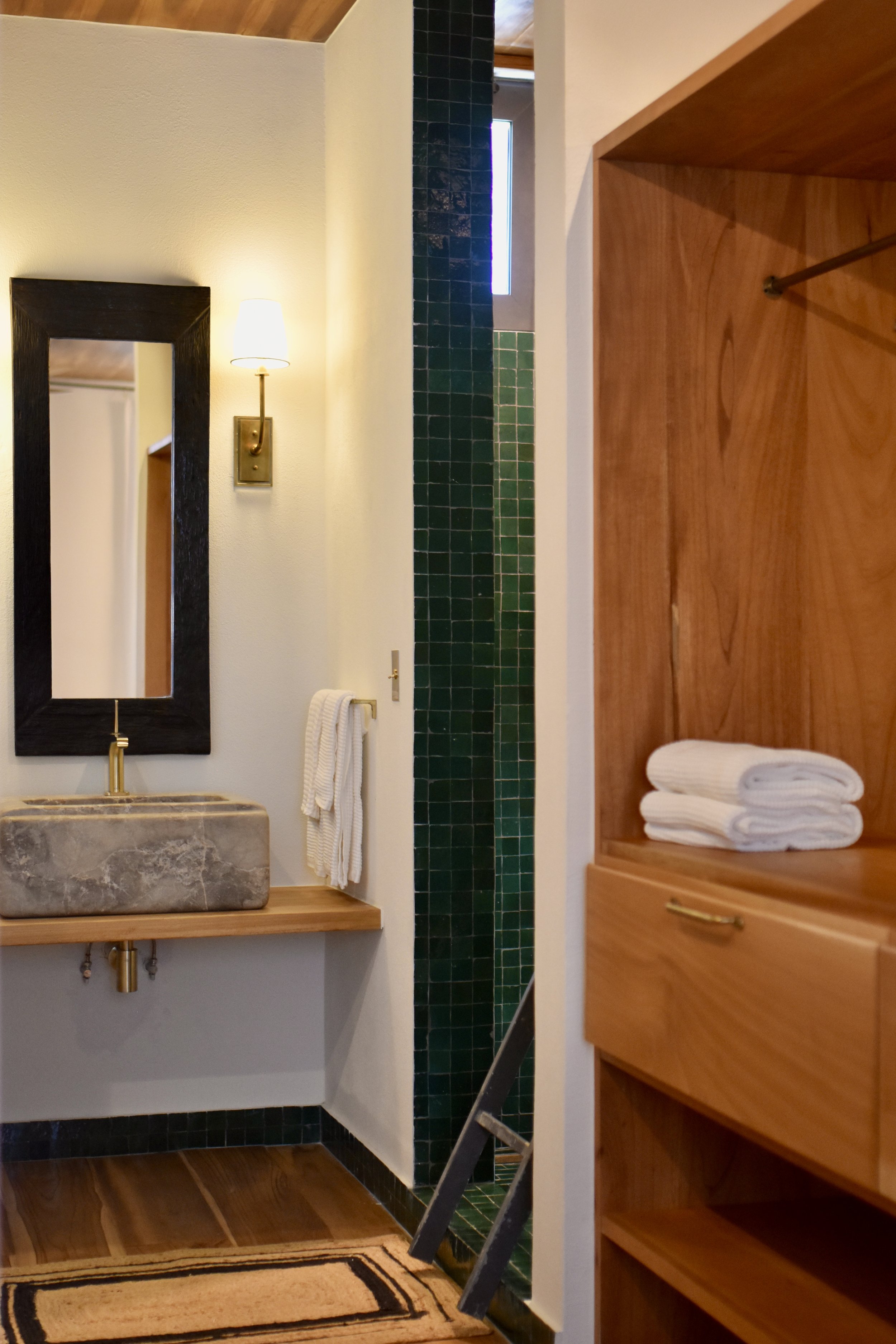 A bathroom with a stone sink, mirror, wall sconce, and a wooden cabinet with towels, with green and black tiled wall and wooden ceiling.
