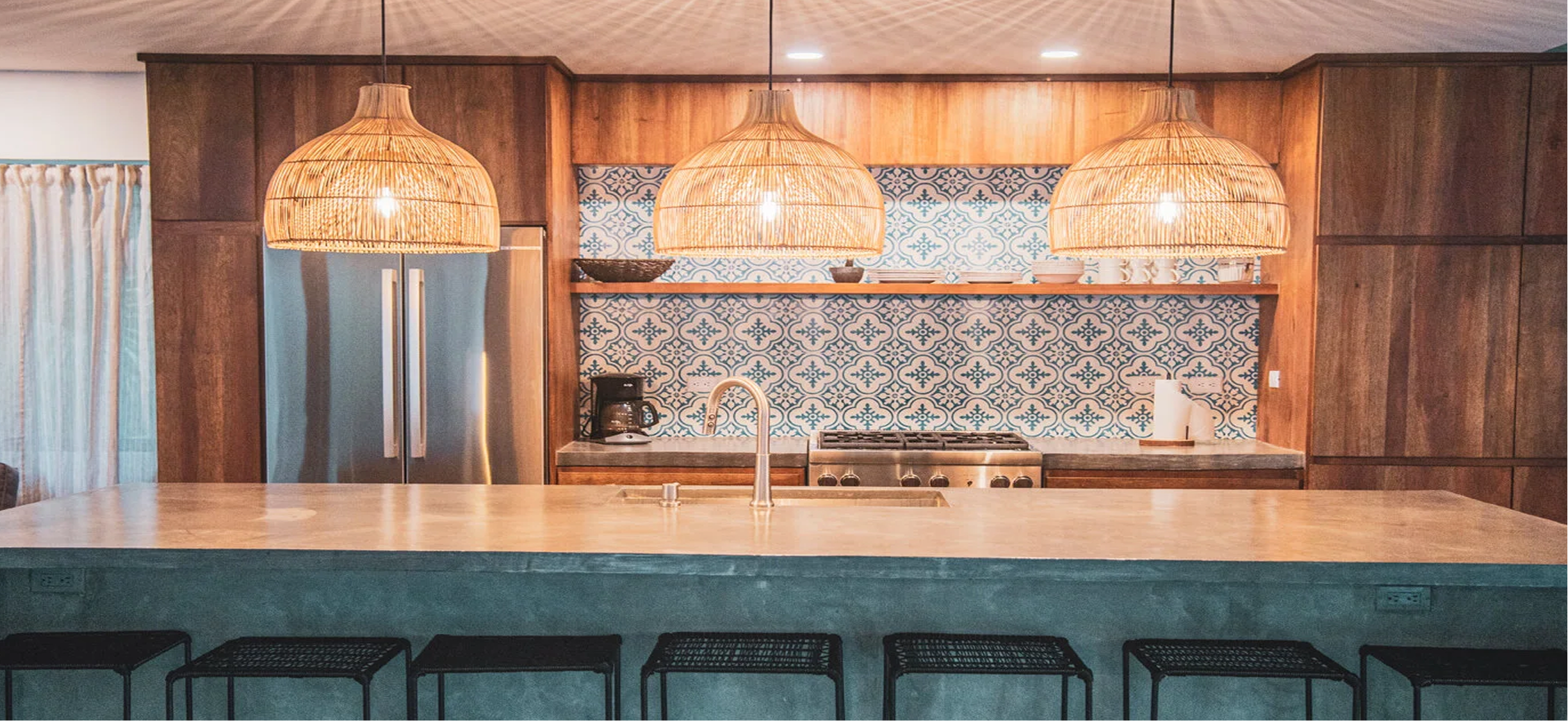 Kitchen with wooden cabinets, blue patterned backsplash, large island with a beige countertop, three wicker pendant lights, stainless steel fridge, coffee maker, and bar stools.