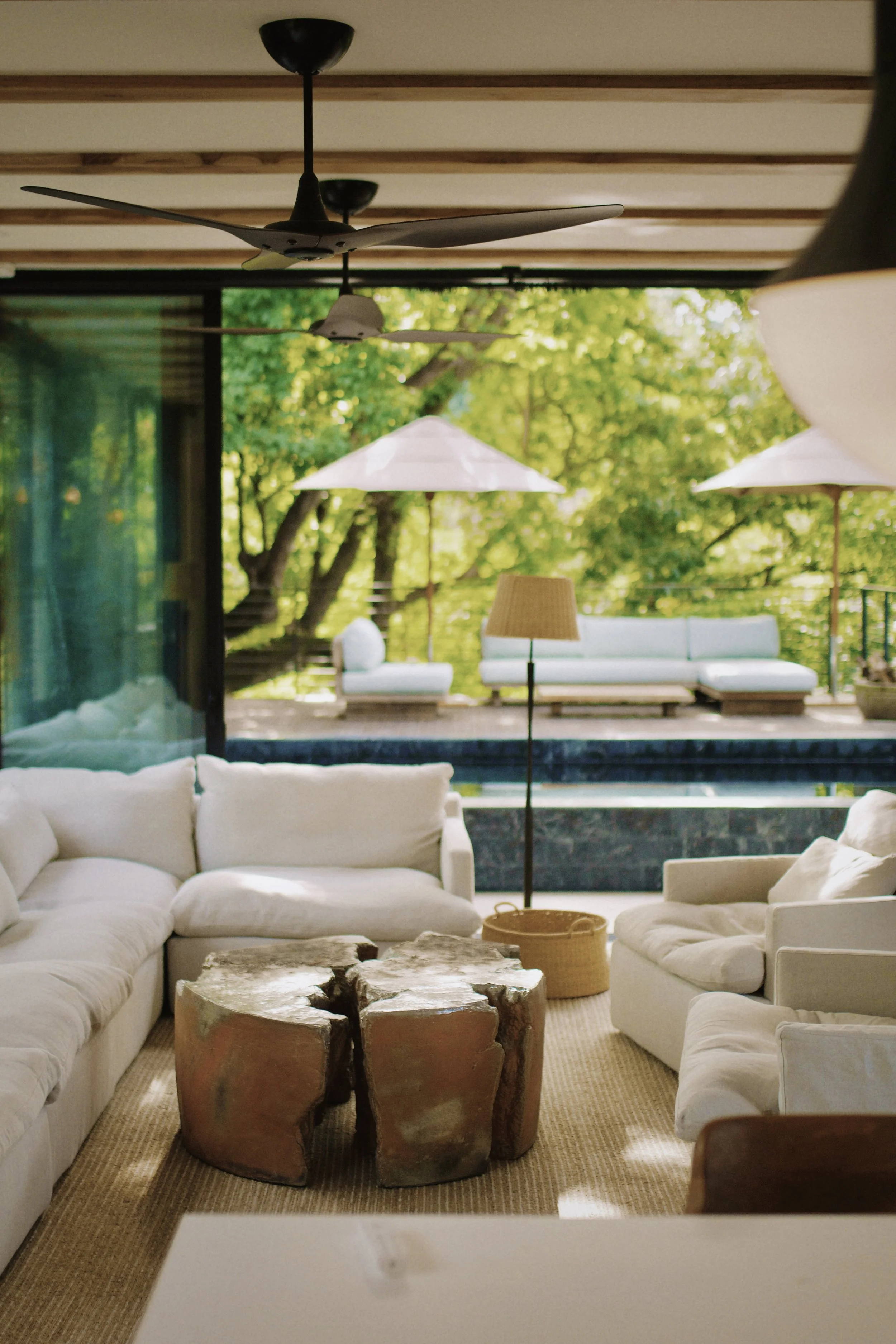 A living room with white sofas, a unique wooden coffee table, and a view of a patio with outdoor seating and lush green trees outside.