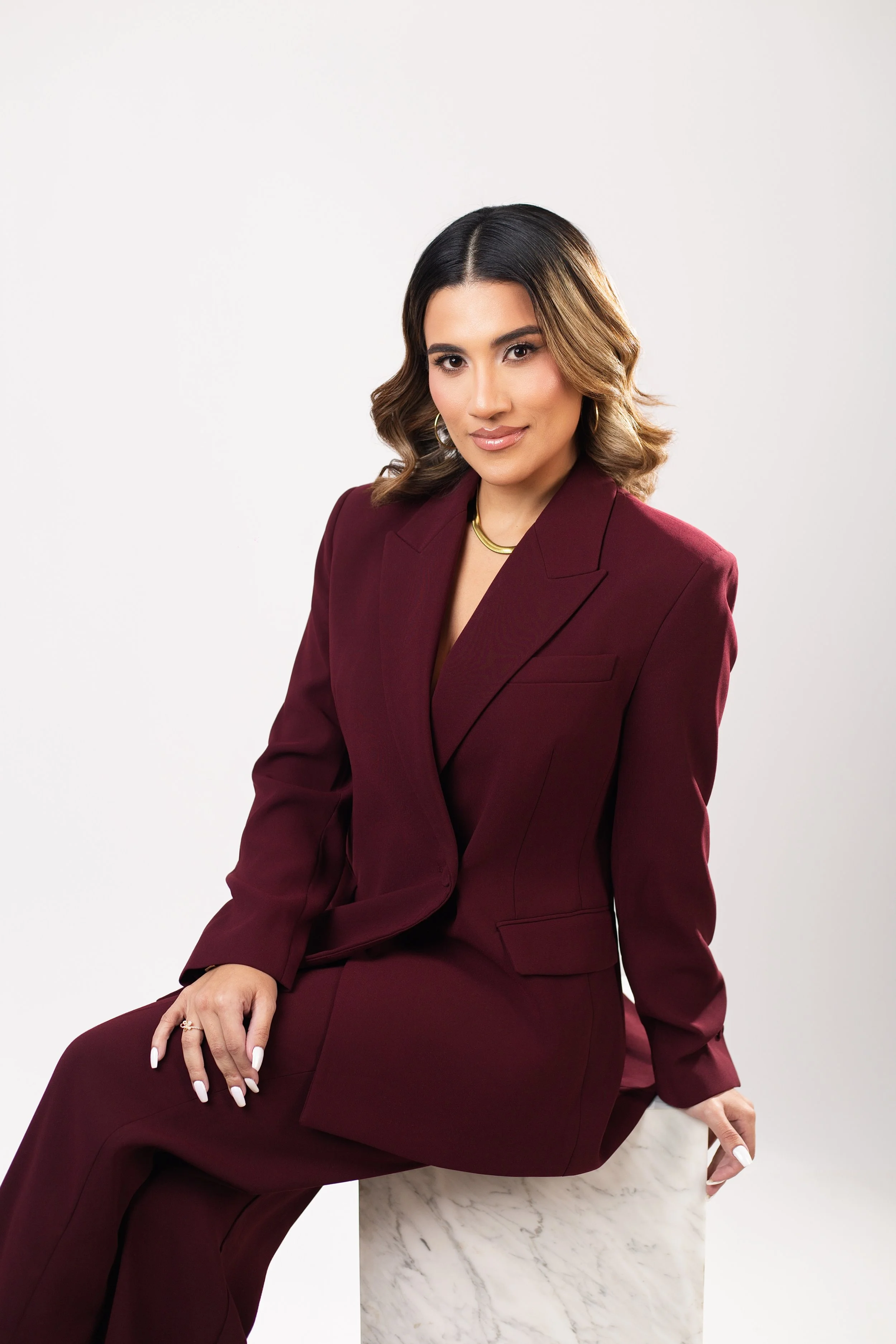 A woman with shoulder-length wavy hair in a dark burgundy suit, sitting on a marble stool against a plain white background.