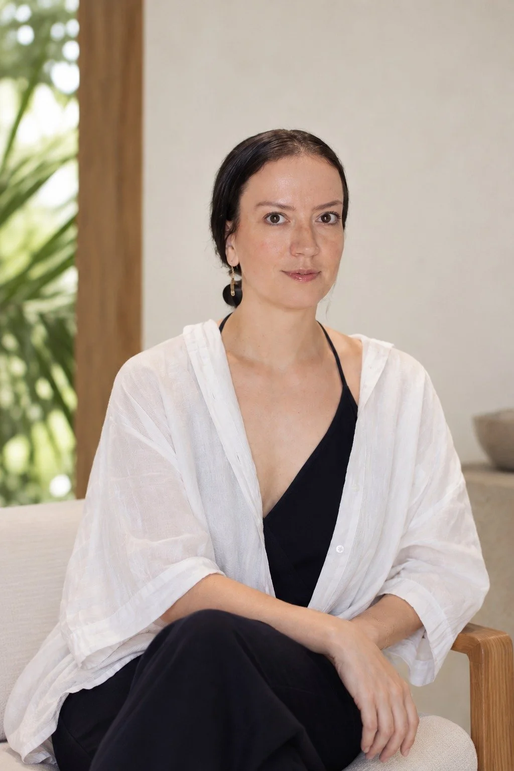 A woman with dark hair tied back, wearing a black top and a white sheer cover-up, sitting on a light-colored chair with a wooden armrest, indoors with green foliage visible through a window.