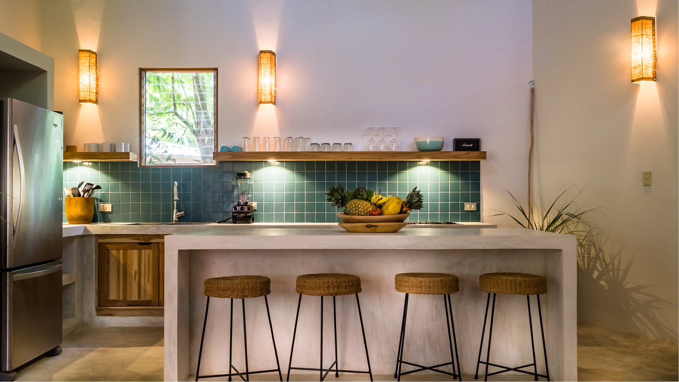 Modern kitchen with a white island, hanging woven bar stools, a fruit bowl, green tiled backsplash, upper open shelf with glasses, and wall-mounted lights.