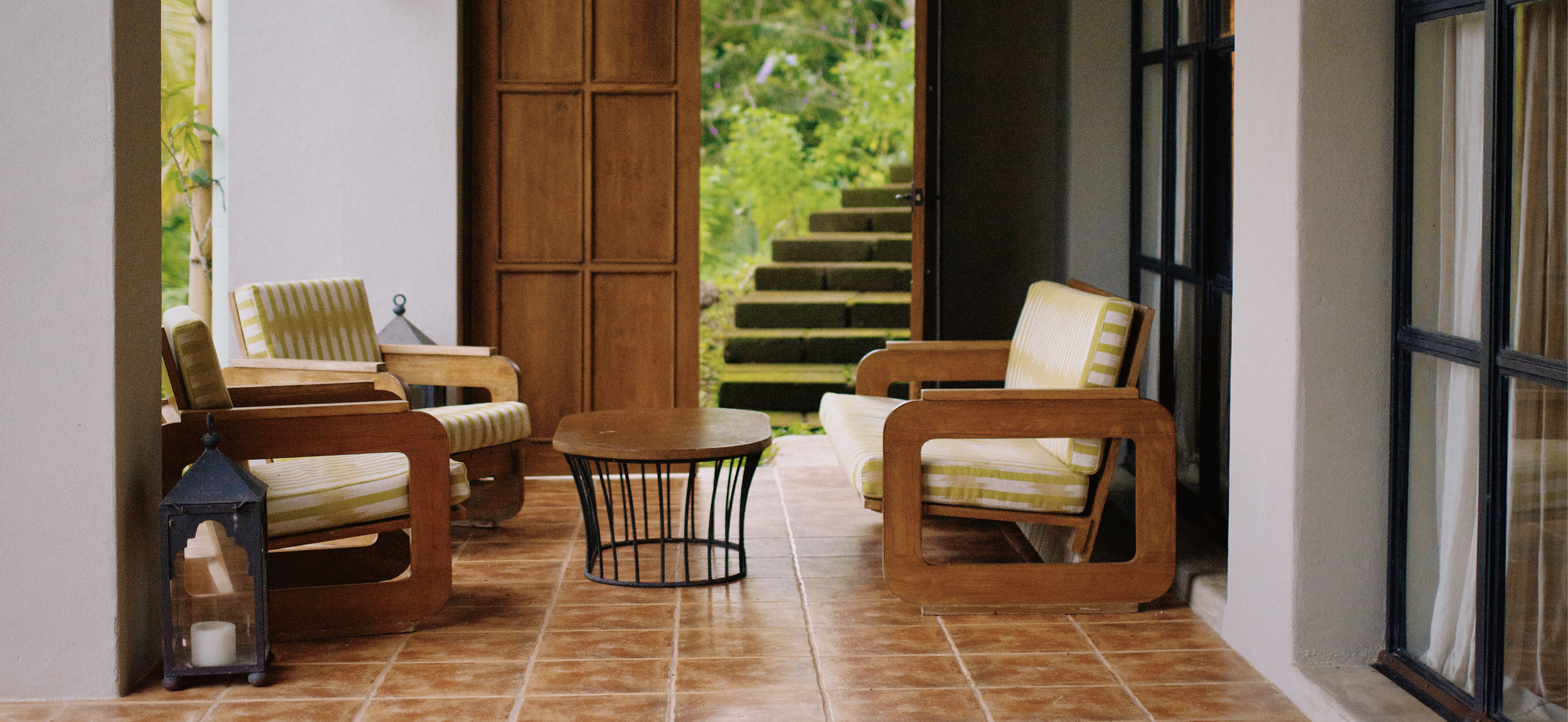 Covered patio with two wooden armchairs with yellow-striped cushions, a round wooden coffee table, and decorative lanterns with candles inside. An open door leads outside to steps and lush greenery.