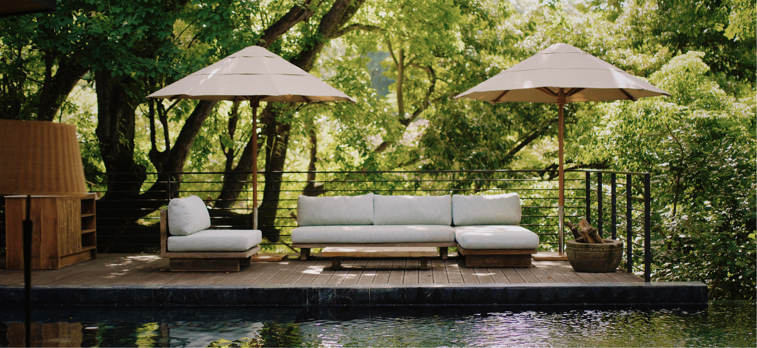 Outdoor patio with white cushioned seating, large beige umbrellas, and lush green trees in the background, overlooking a body of water.