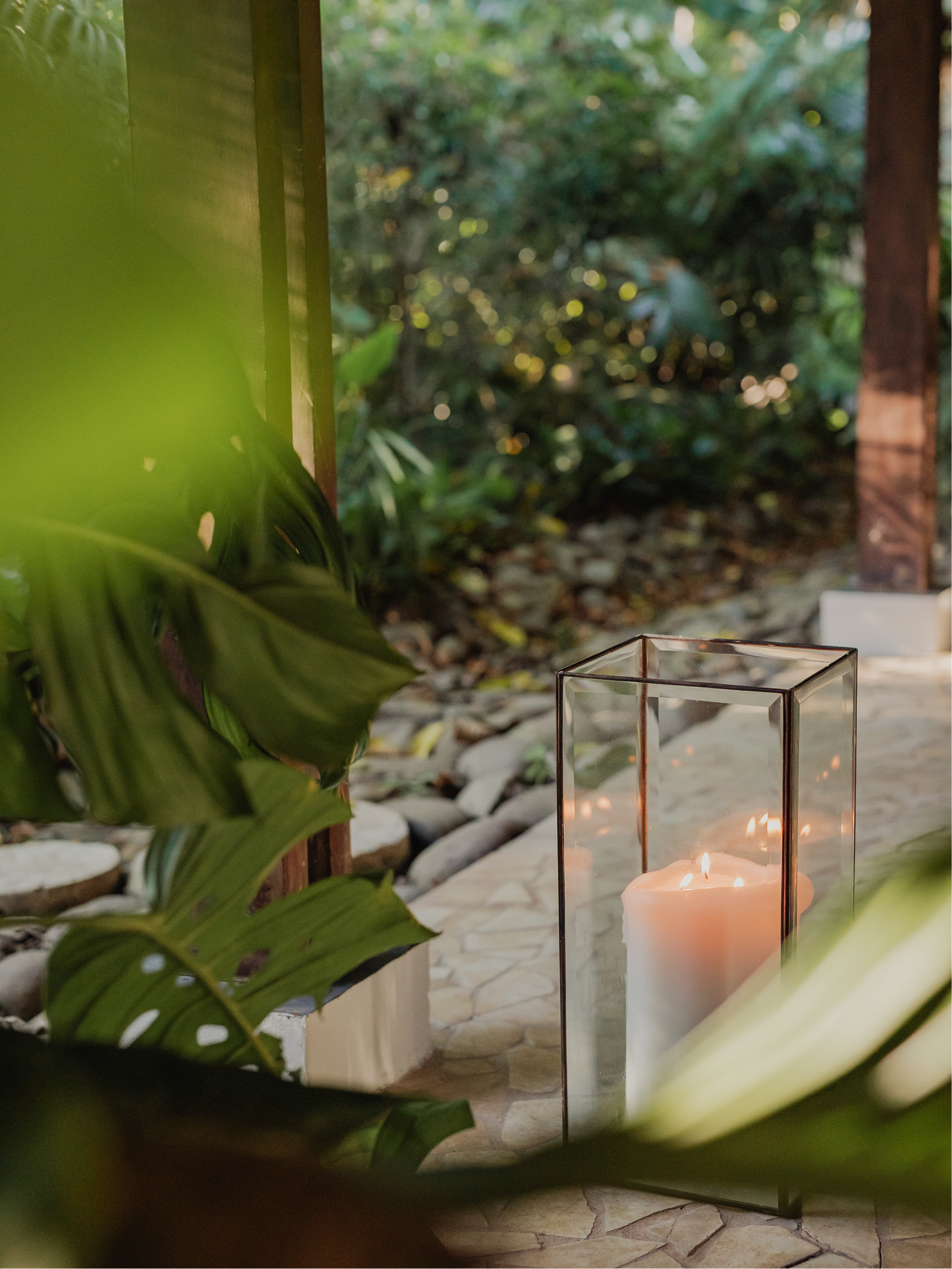 A lit white pillar candle inside a clear glass lantern with a metal frame, placed outdoors on stone paving, with lush green foliage and trees in the background.