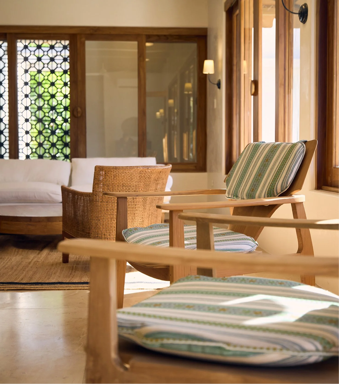 A cozy sitting area with wooden furniture, including armchairs with striped cushions and a white sofa, illuminated by natural light through large wooden-framed windows, with decorative wall lamps on a beige wall.