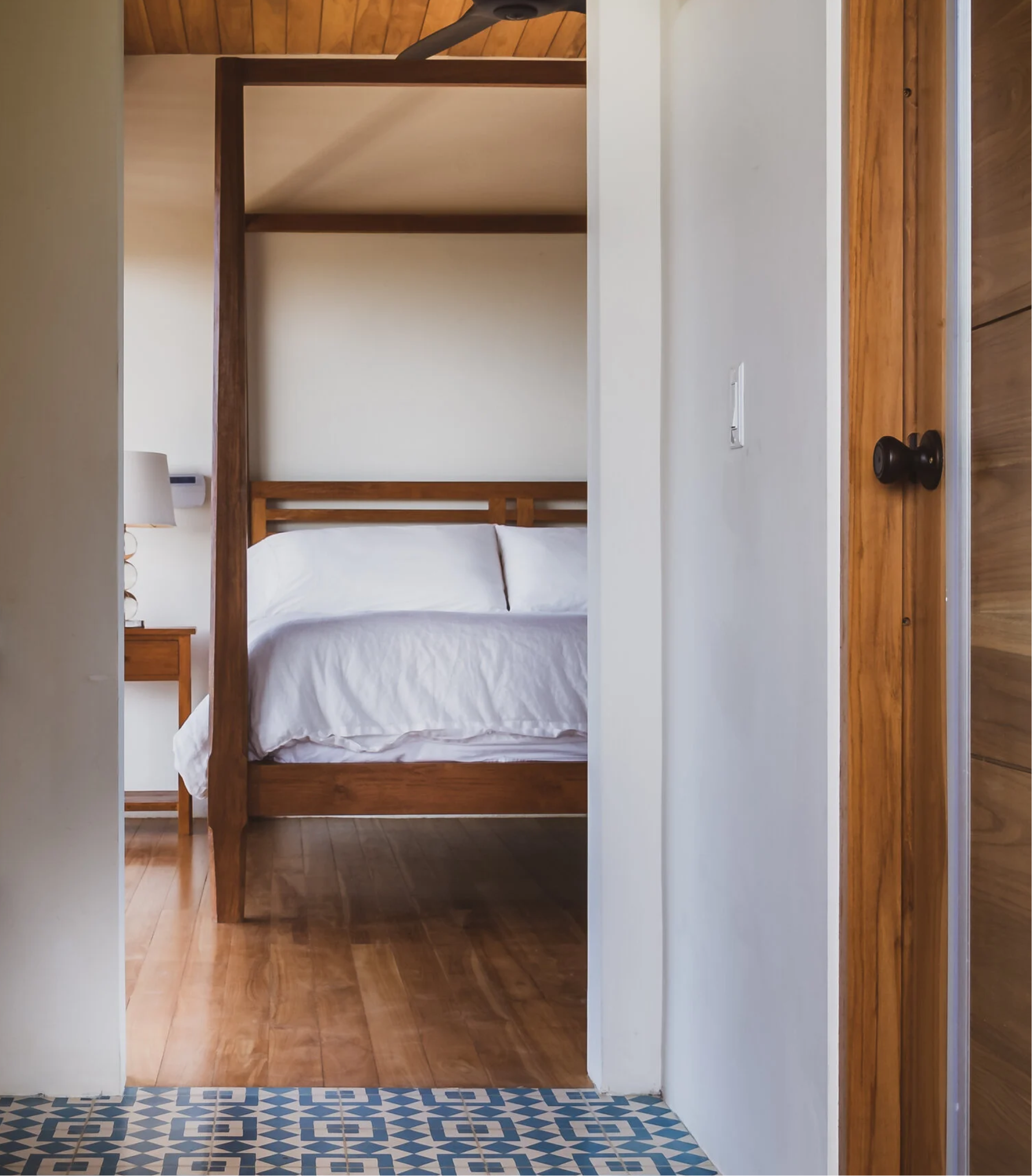 View of an interior of a room with a wooden bed frame, white walls, a wooden ceiling, a small bedside table, a white lamp, a white blanket on the bed, and a window with shutters.