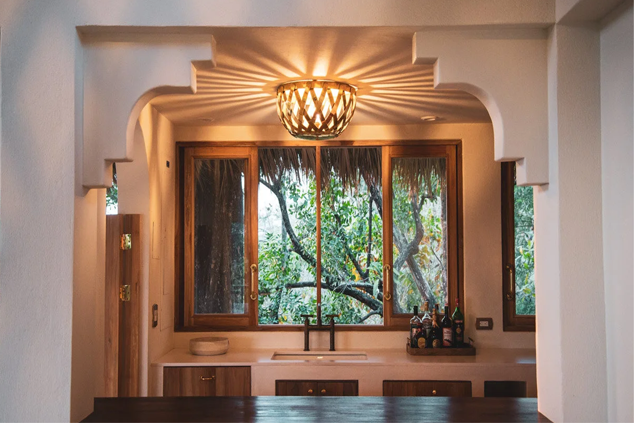 Interior view of a room with a large window showing trees outside. The ceiling has a decorative light fixture casting shadows on the ceiling. Below the window, some bottles are placed on a surface.