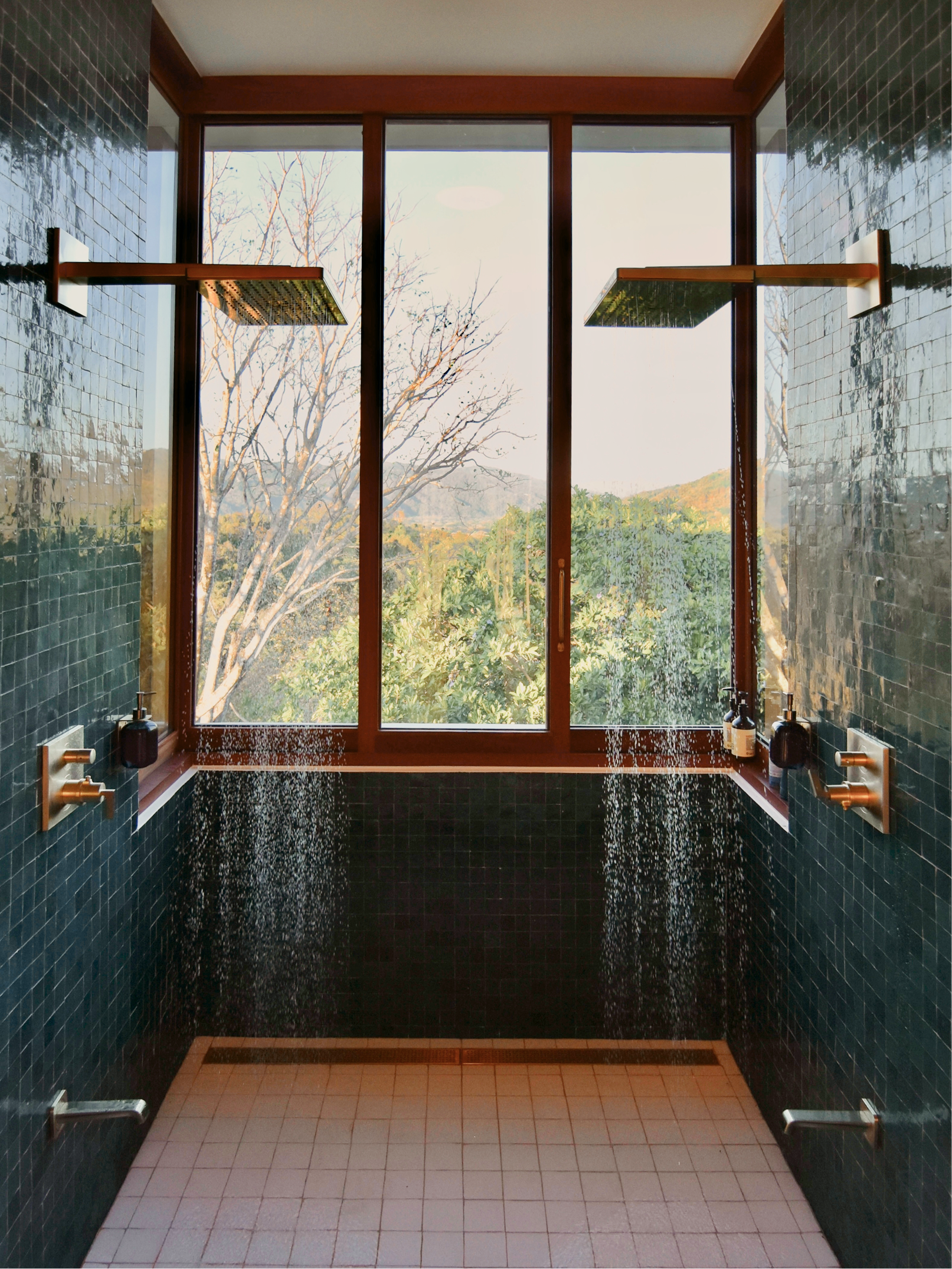 Indoor shower with two large shower heads, dark green tiled walls, large window showing trees outside, and white tiled floor.