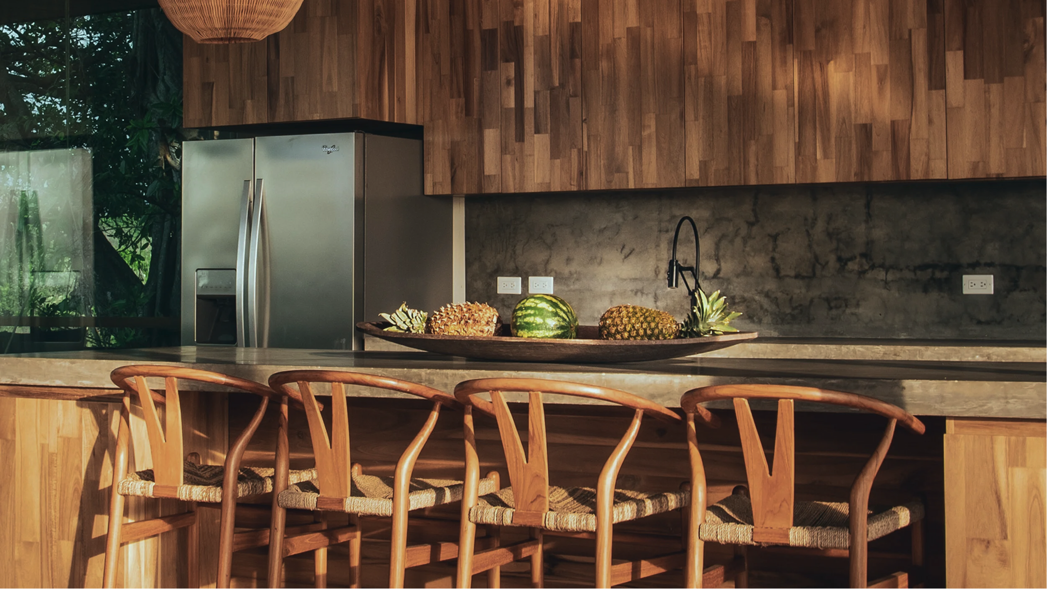 Modern kitchen with wooden cabinets, a concrete backsplash, a stainless steel refrigerator, and a large bowl of tropical fruits including pineapple, watermelon, and pineapple on a black tray. Wooden chairs with woven seats are in the foreground.