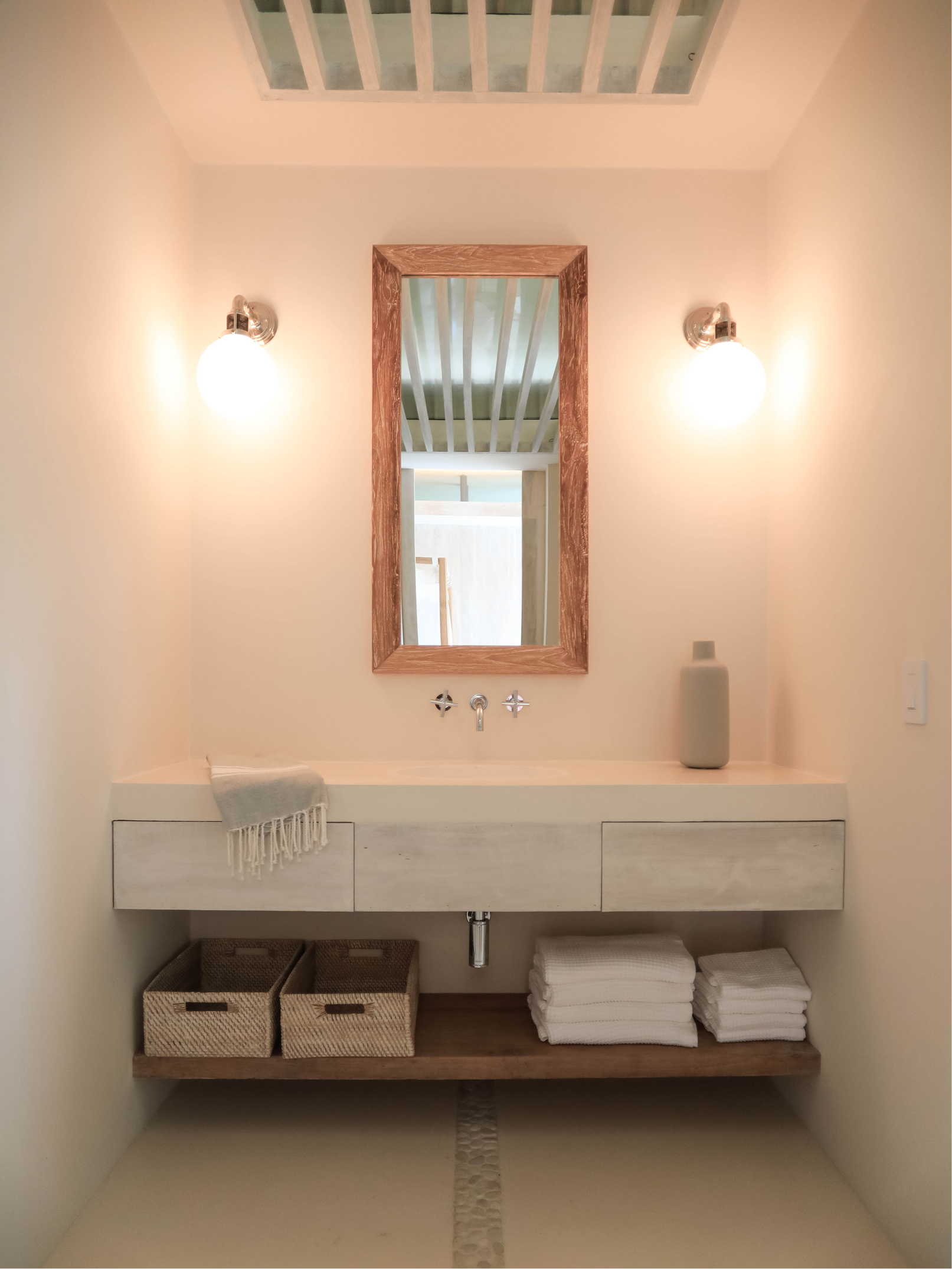 A bathroom vanity with a white countertop, a rectangular mirror with a wooden frame, wall-mounted light fixtures, a gray ceramic vase, three folded white towels, and two wicker baskets on a wood shelf. The floor has a pebble-stone drainage channel.