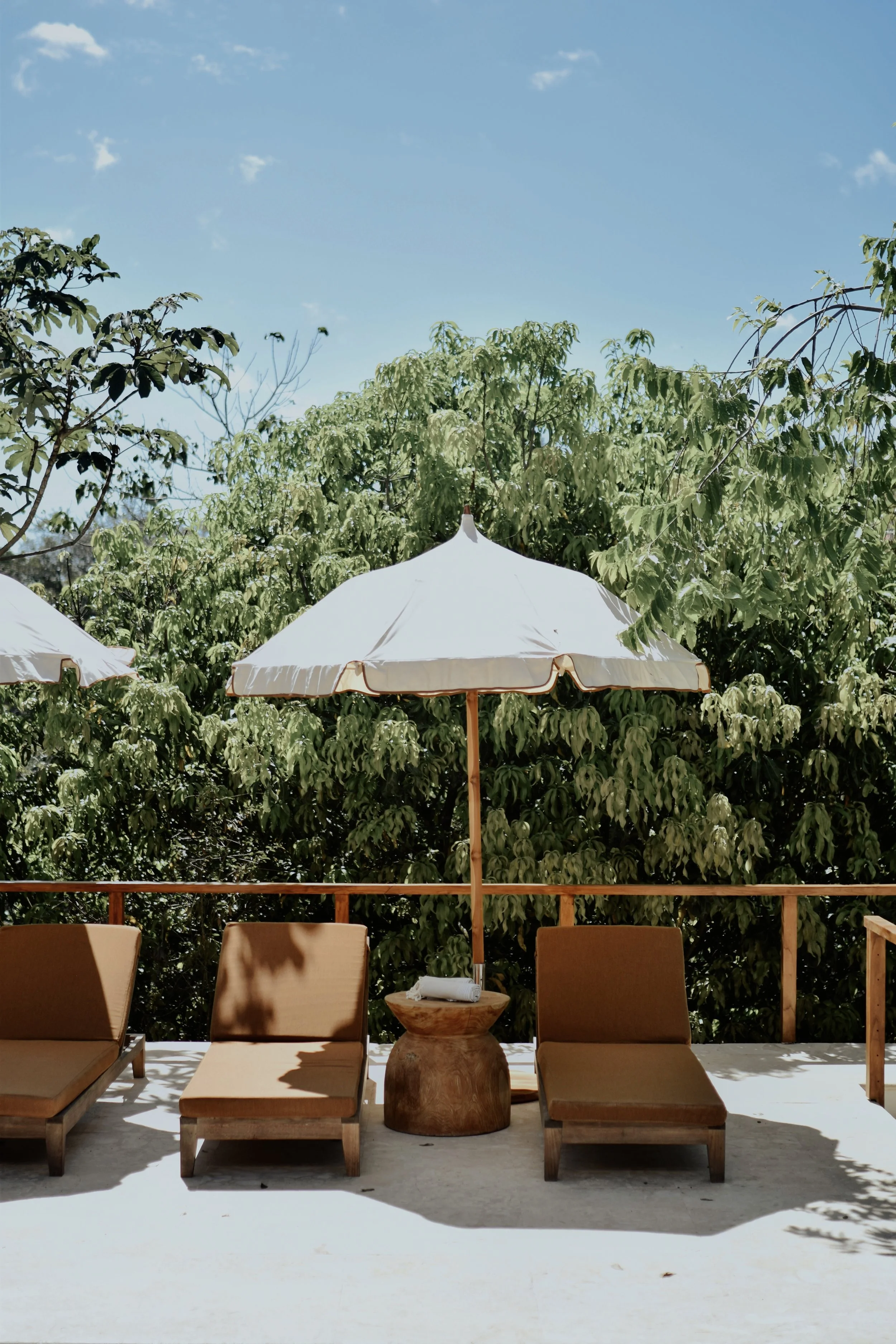 Outdoor patio with four brown cushioned lounge chairs, a small wooden side table with a rolled towel on it, and two large white patio umbrellas with wooden poles. Green trees and a clear blue sky in the background.