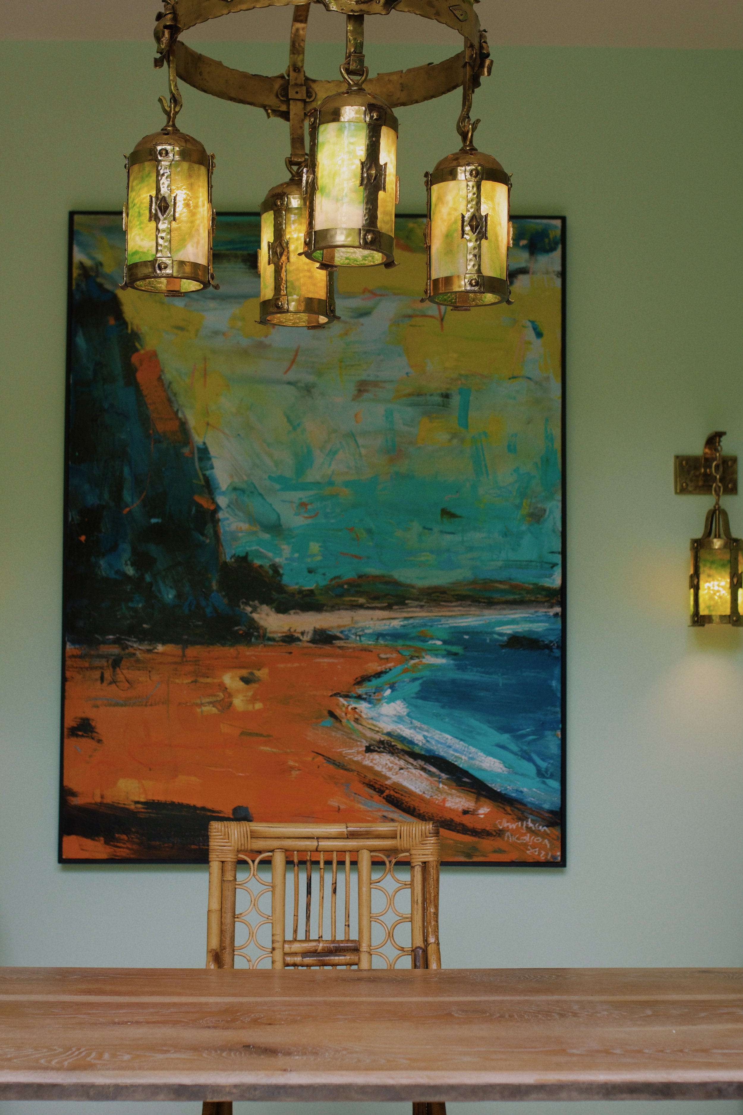 A wooden table with a rattan chair in front of an abstract landscape painting on a light green wall. Hanging from the ceiling is a vintage lantern-style chandelier with five colorful glass shades.